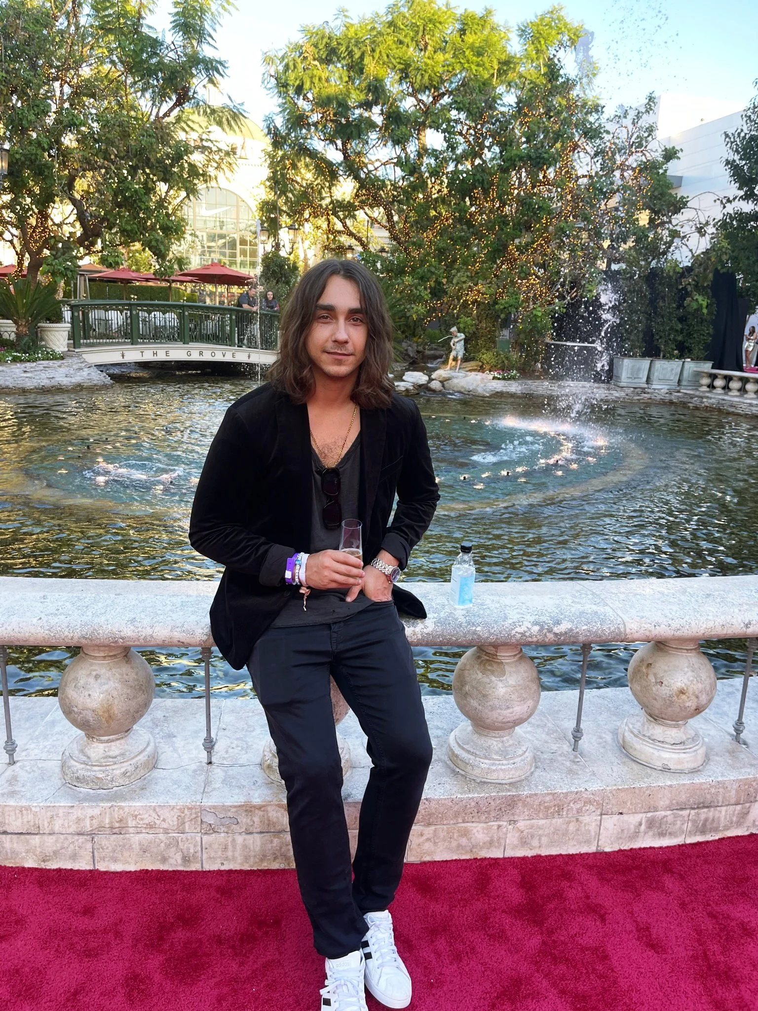 A young native american man named matthew blocha with long brown hair standing on a red carpet near a fountain with trees and a bridge in the background, holding a glass of drink.