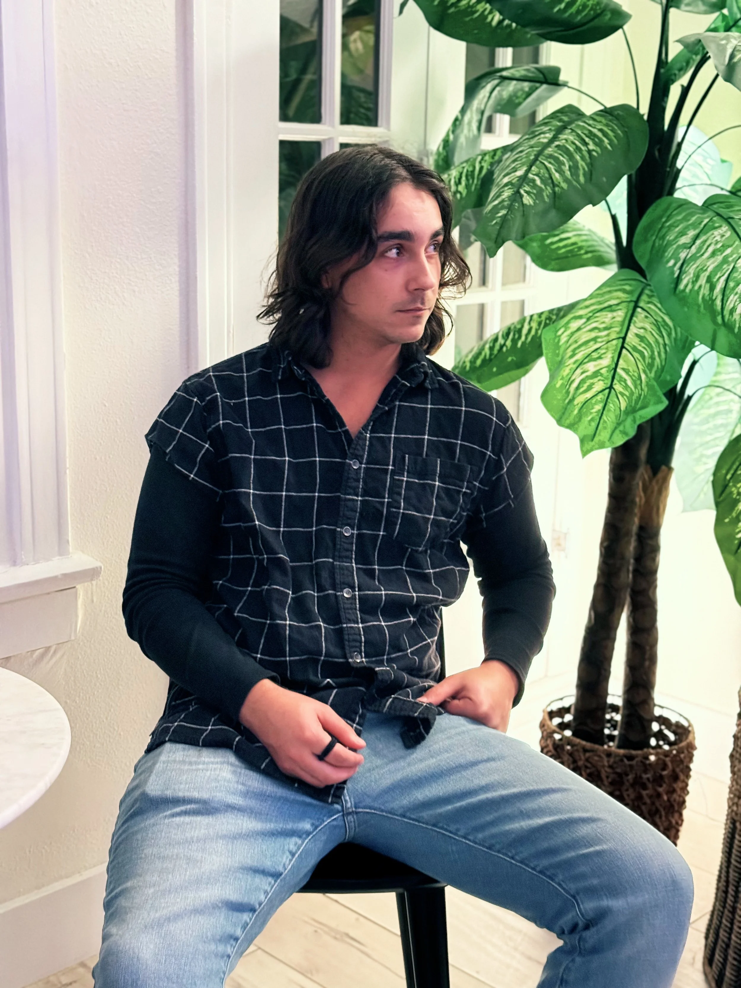 A young man with shoulder-length dark hair and light skin, wearing a black and white checkered shirt over a black long-sleeve shirt, seated on a black stool next to a large green plant with variegated leaves, in a well-lit indoor space with white wal