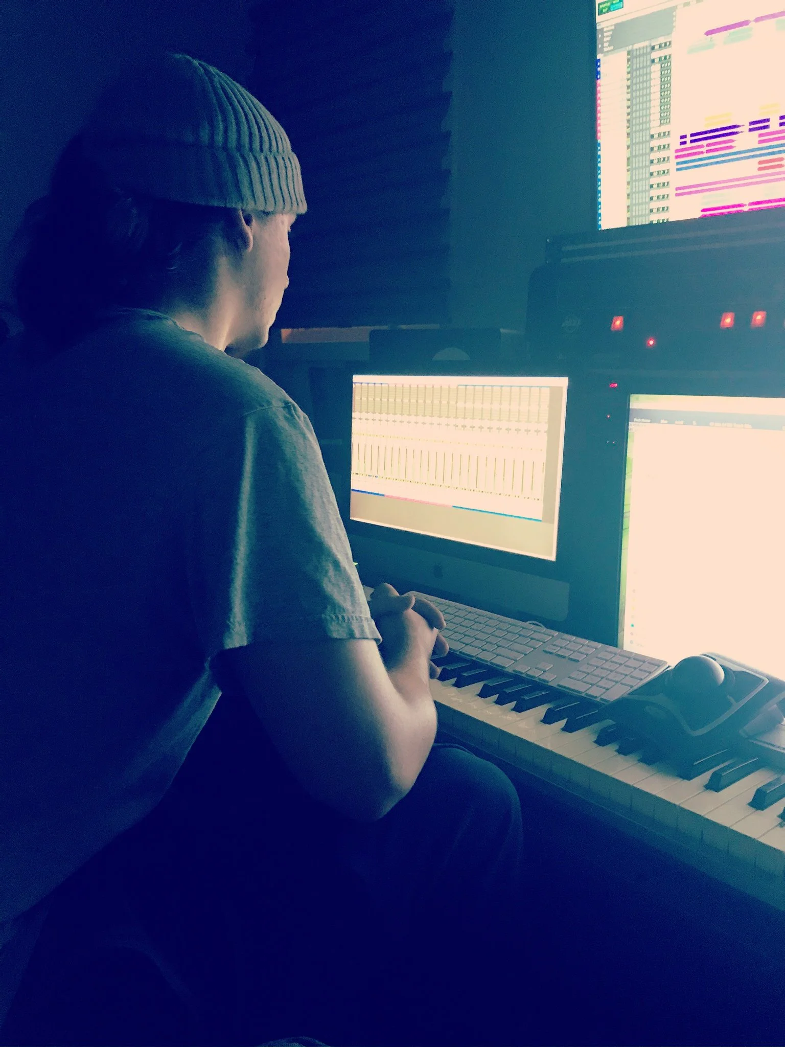 Person wearing a beanie working on music production in a dimly lit room with multiple monitors displaying music software and a keyboard.