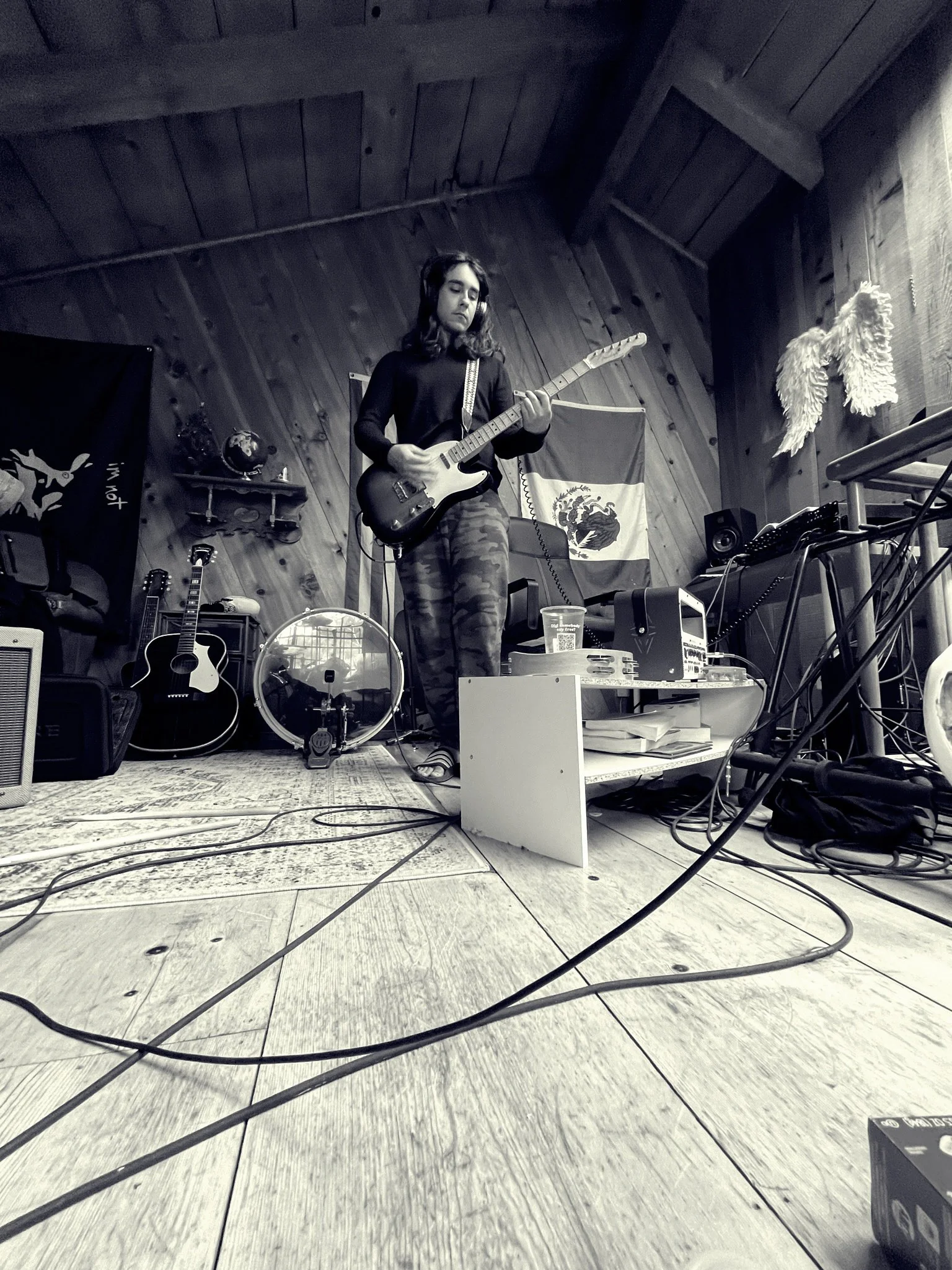 A native american musician named matthew blocha standing on a wooden floor, playing an electric guitar in a rustic room with wood-paneled walls. There are musical instruments such as guitars, a drum, and an amplifier around him