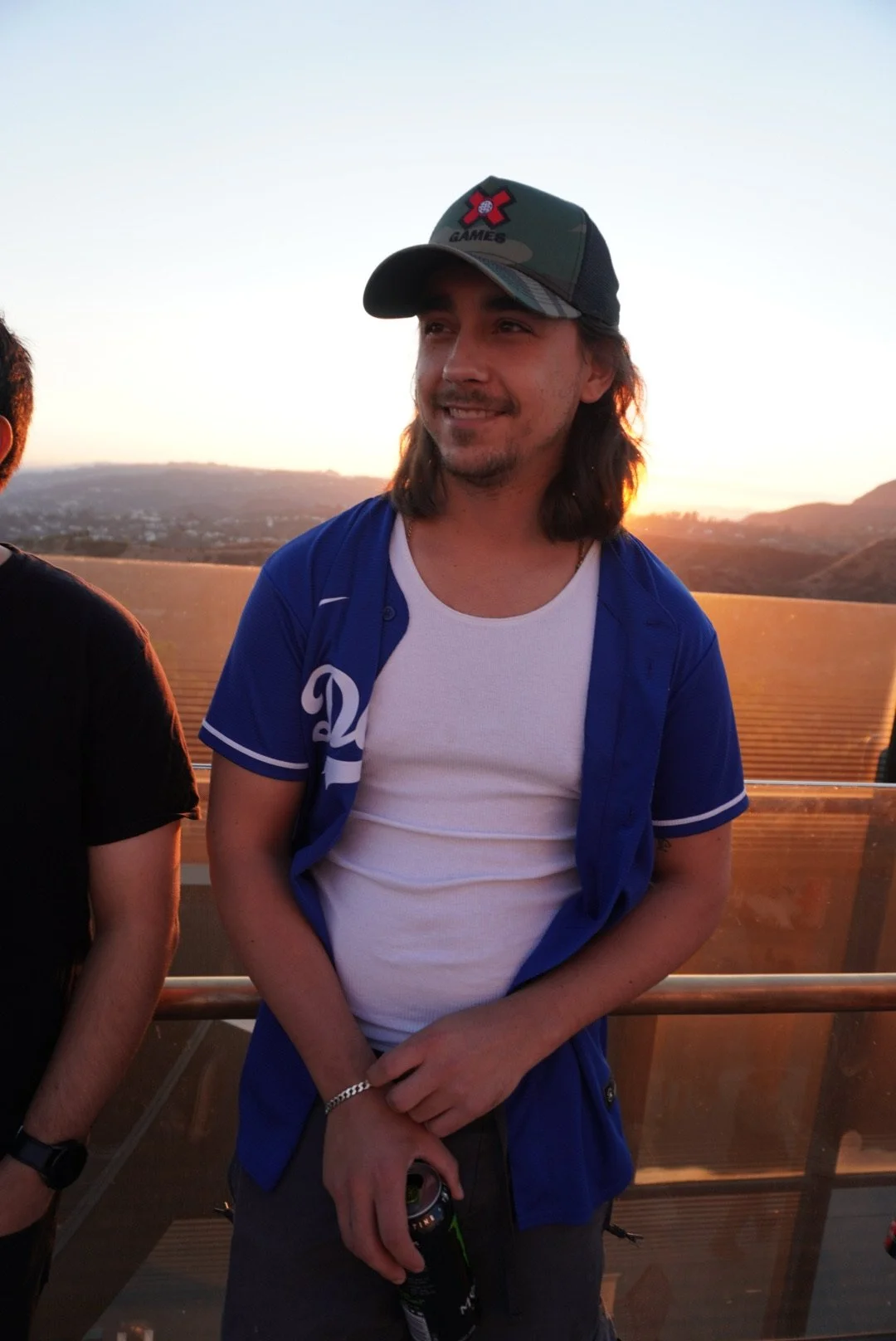 A native american man, matthew blocha with long hair wearing a baseball cap, a white t-shirt, and a blue shirt stands outdoors during sunset, smiling and holding a drink.