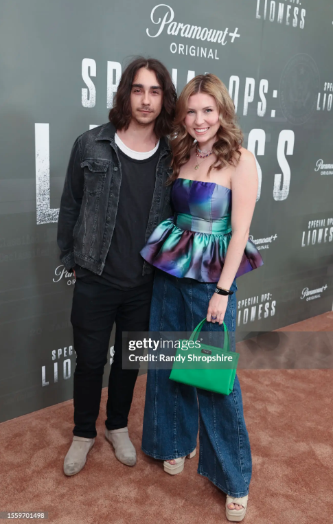 Matt blocha, a native american man with shoulder-length curly hair and a woman with blonde curly hair, both smiling and posing together at an event. The man wears a black denim jacket over a black shirt and black pants. The woman wears colors