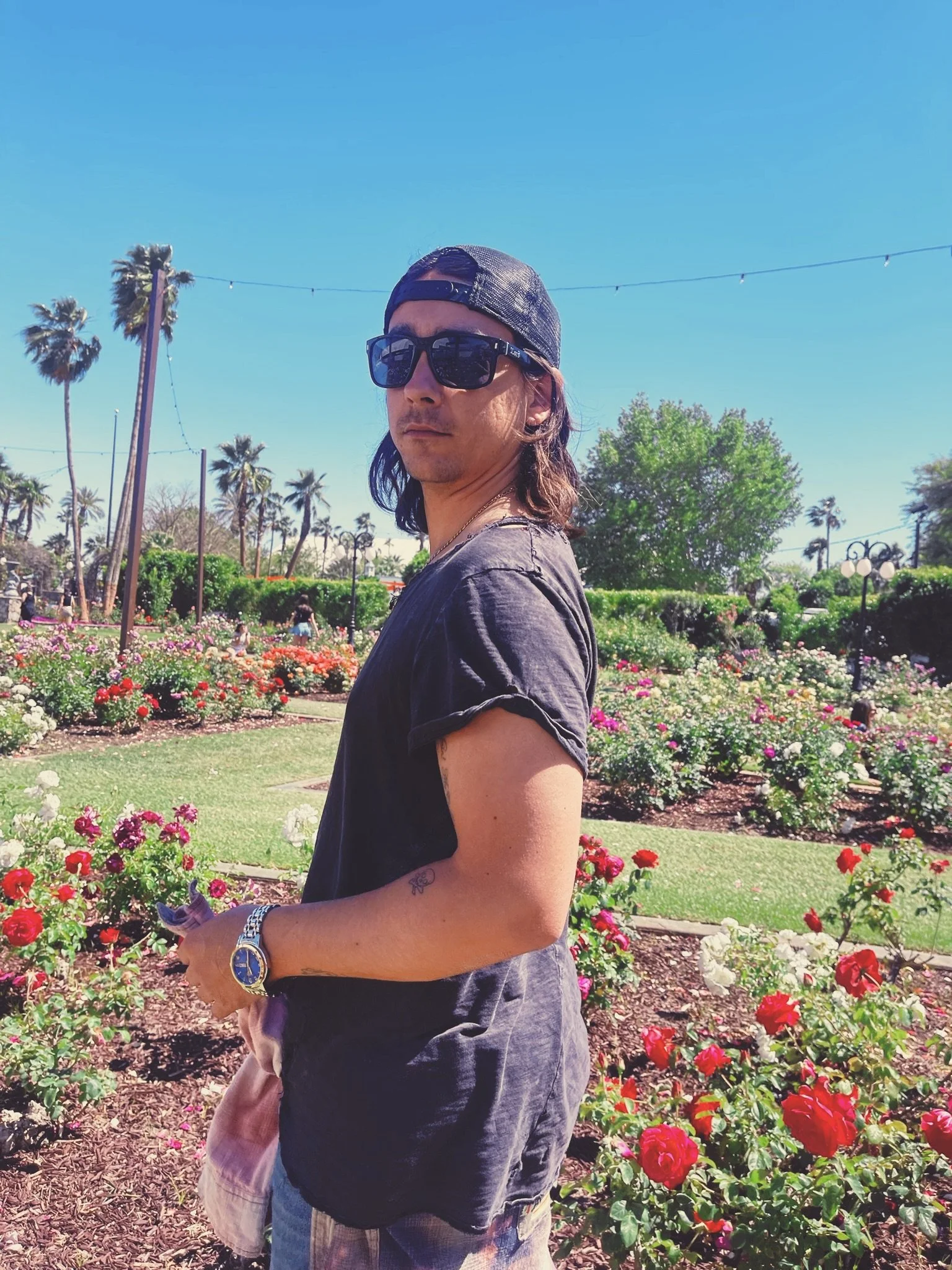 native american Young man named matthew blocha with long hair, wearing sunglasses, a backwards cap, black t-shirt, and watch, standing outdoors in a garden with roses, palm trees, and a clear blue sky in the background.