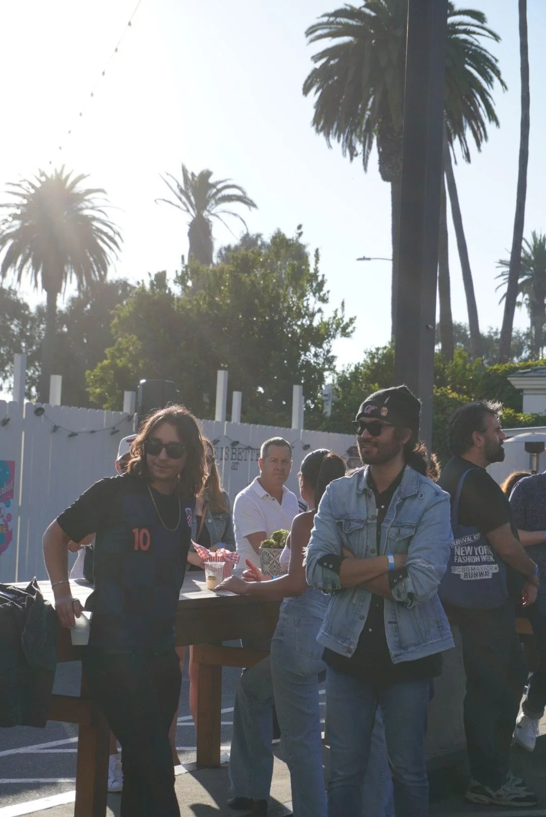 Matt Blocha and Flight By Nothing's bassist Porfi Rubio gathered outdoors at a casual event, some wearing sunglasses and standing near a high table with food and drinks, with palm trees and string lights in the background.