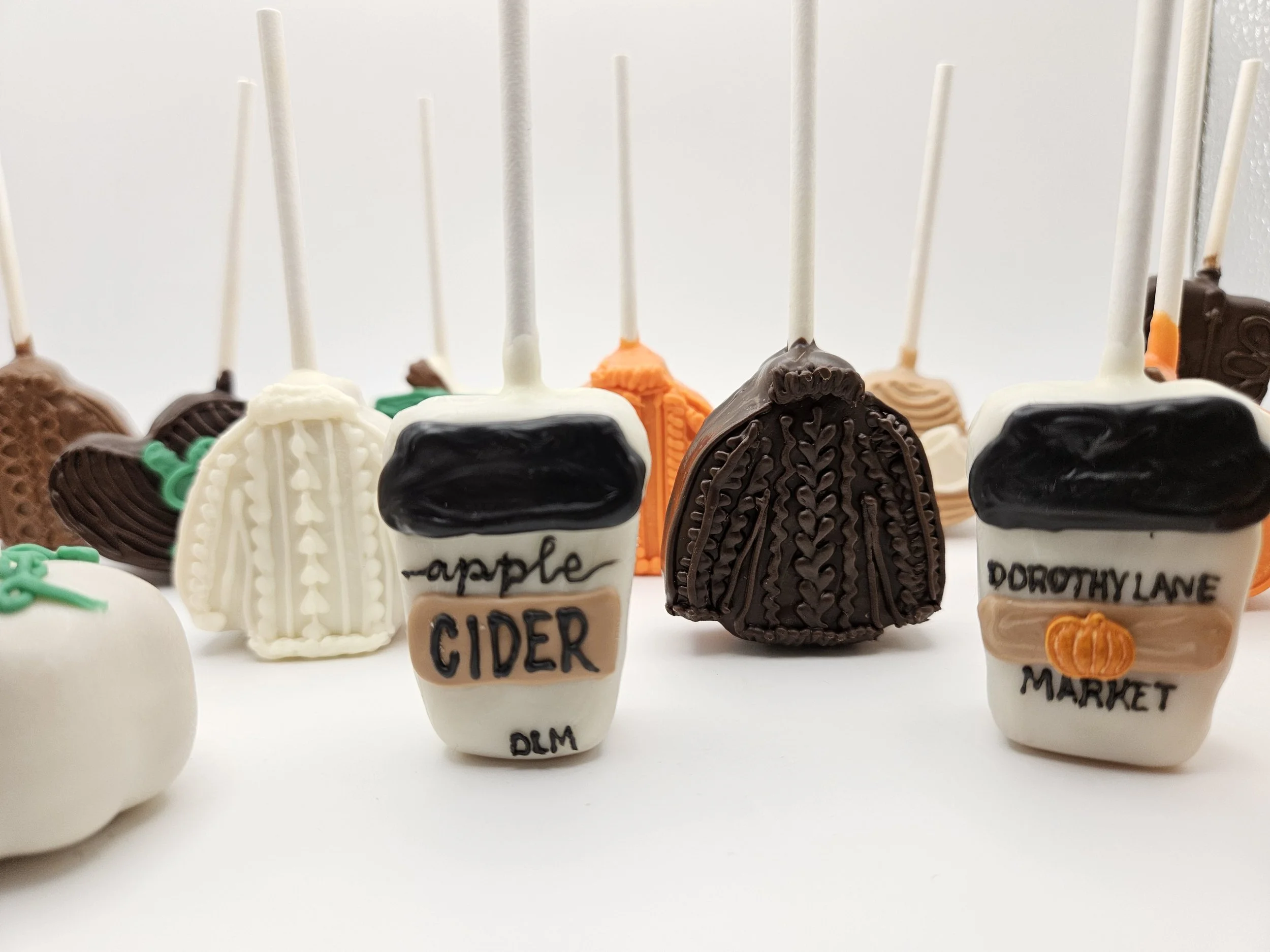 Decorative cake pops shaped like to-go coffee cups and pumpkins, with some labeled 'Apple Cider' and 'Dorothy Lane Market', arranged on a white surface.