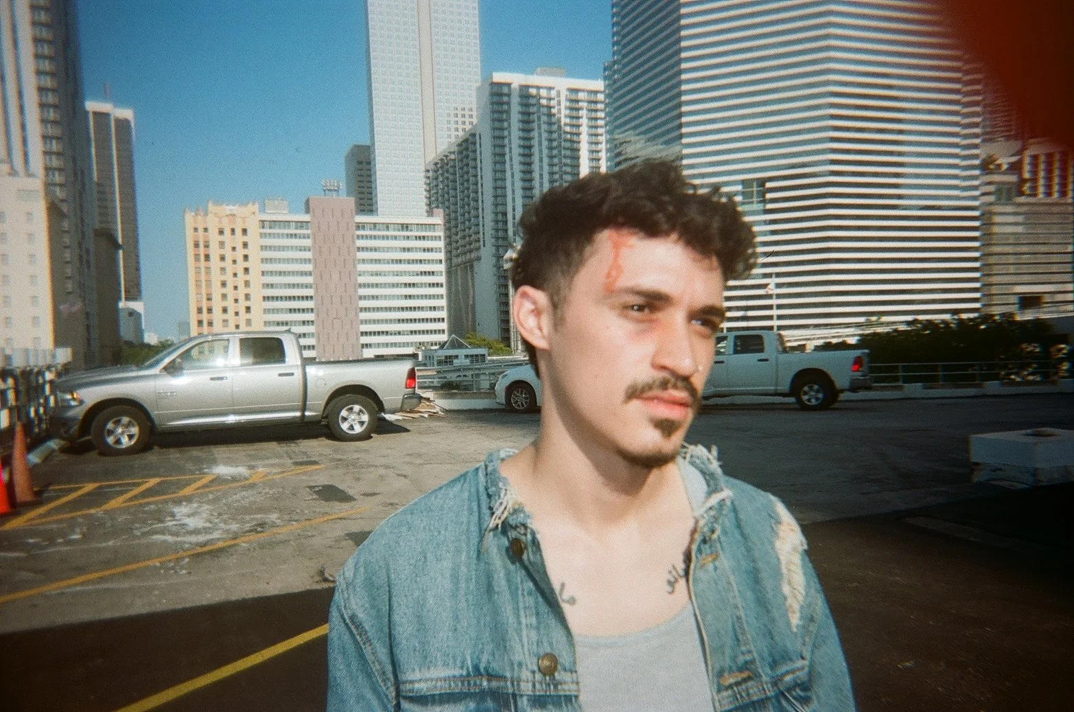A young man with dark curly hair, a mustache, and a small goatee, standing on a rooftop parking lot in an urban area with tall buildings in the background. He is wearing a distressed denim jacket over a gray shirt and has a scratch on his forehead.