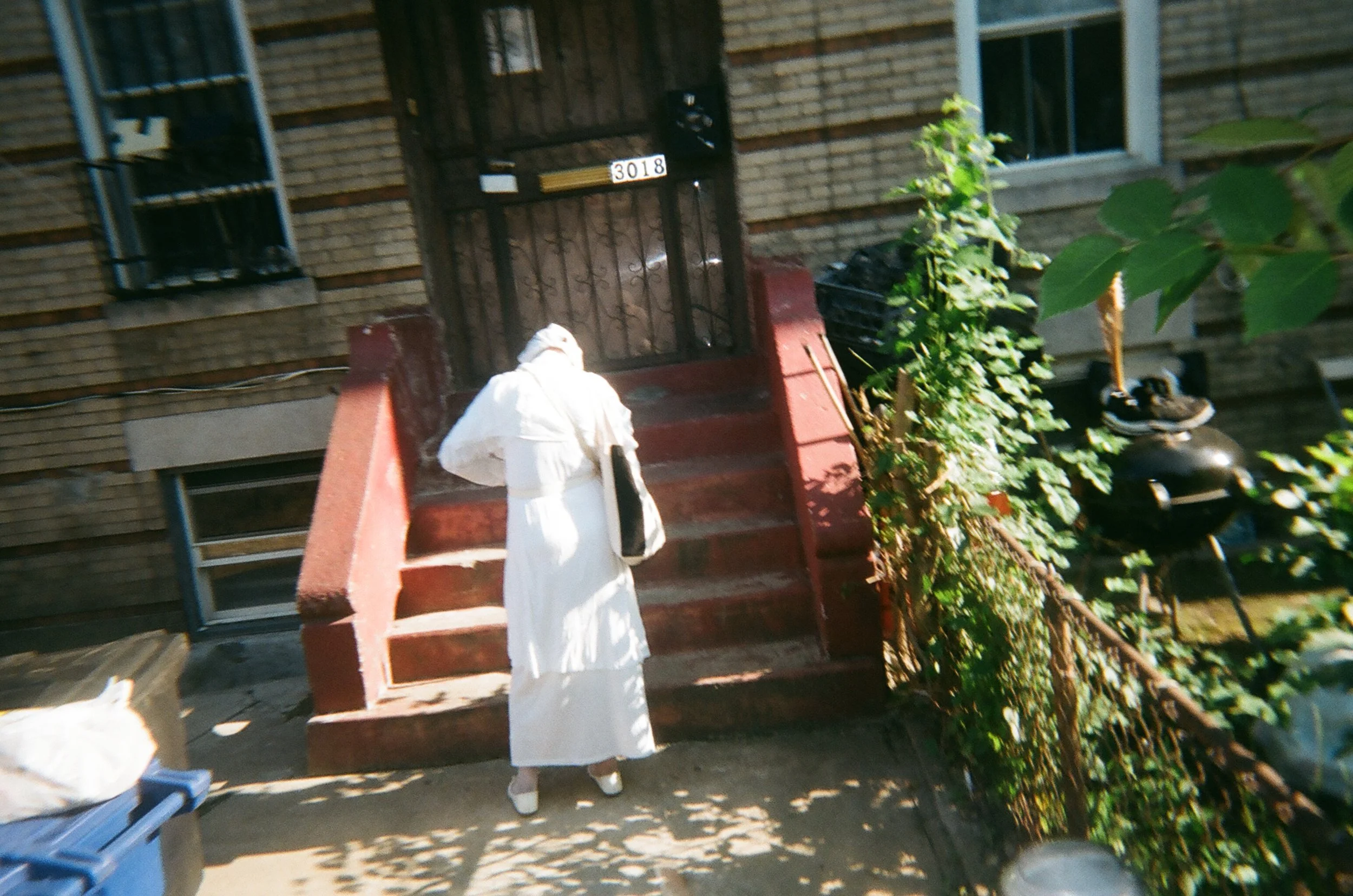 A person dressed in white standing on the bottom of a staircase outside a brick building, knocking on the door. The door has the number '3018' and the stairs are red. There's greenery on the right side and a black grill on the door.