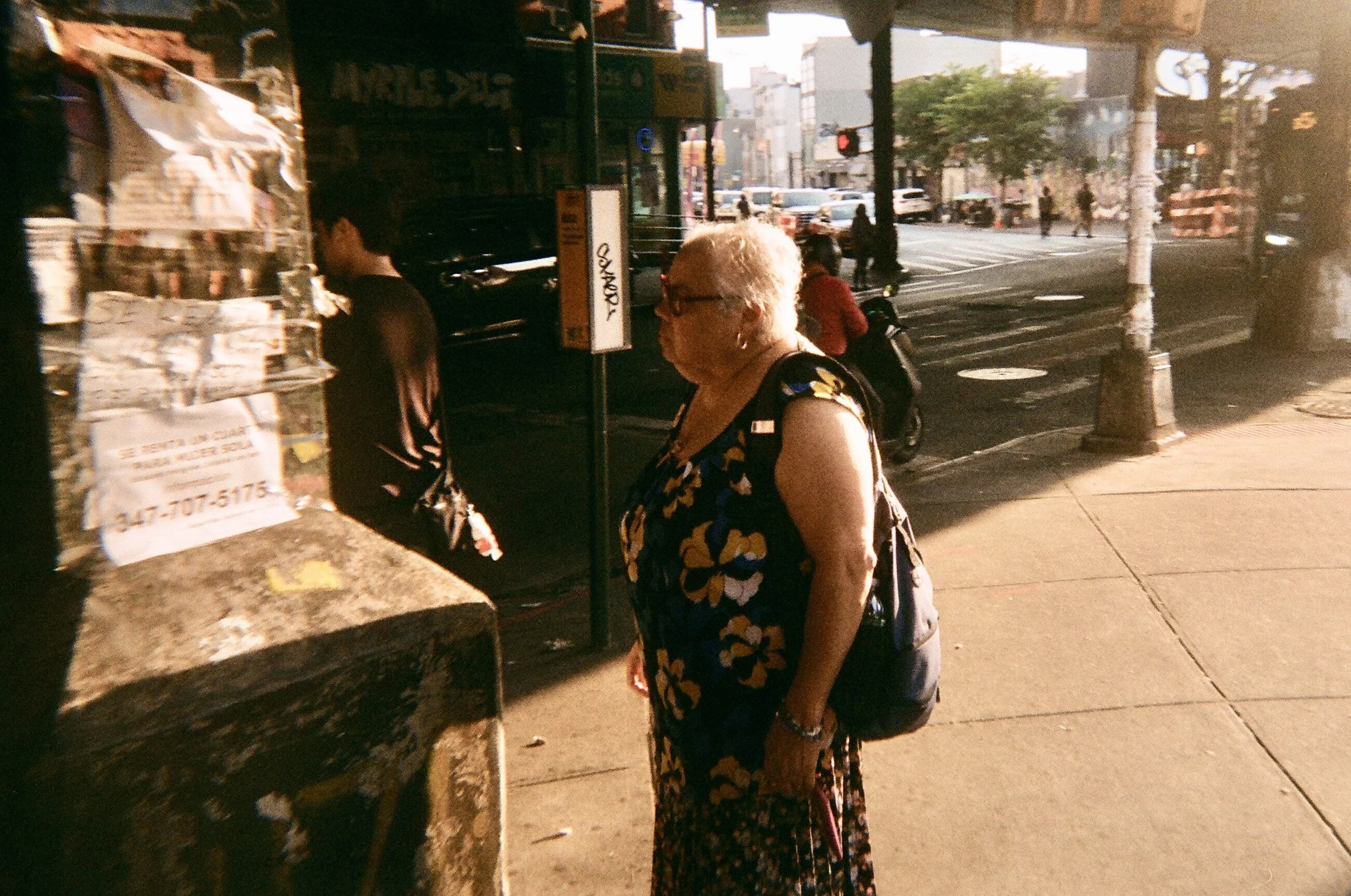 An elderly woman with short white hair, wearing glasses, a sleeveless floral dress, and a backpack, standing on a city sidewalk near a newspaper stand during sunset.