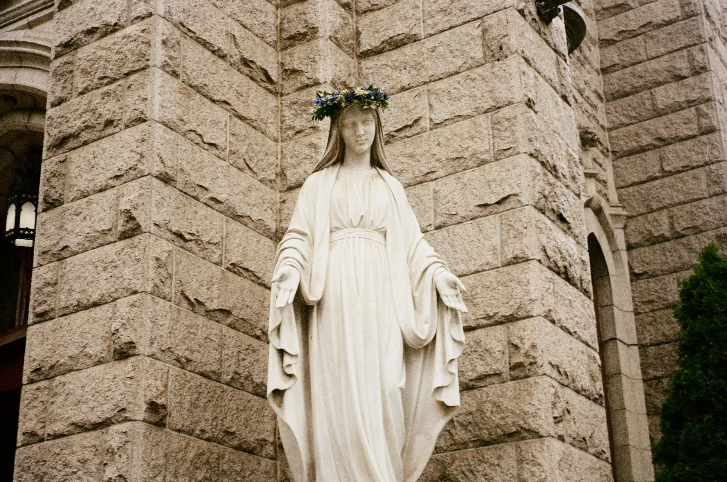 Religious statue of the Virgin Mary with a floral crown, standing outside a stone building.