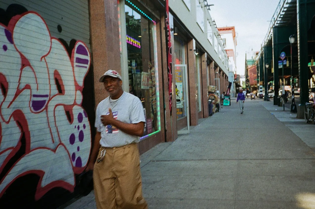 A man standing on a city sidewalk near storefronts with graffiti art on the wall, people walking in the background, and street activity on a cloudy day.