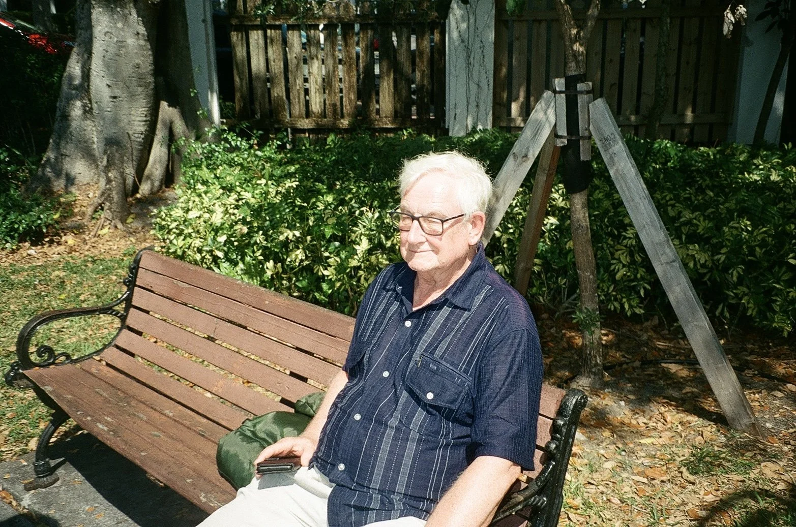 An elderly man with glasses and white hair sitting on a wooden park bench in a garden. He's wearing a dark striped shirt and light-colored pants, with a green bag resting beside him. The garden has trees, bushes, and a wooden fence in the background.
