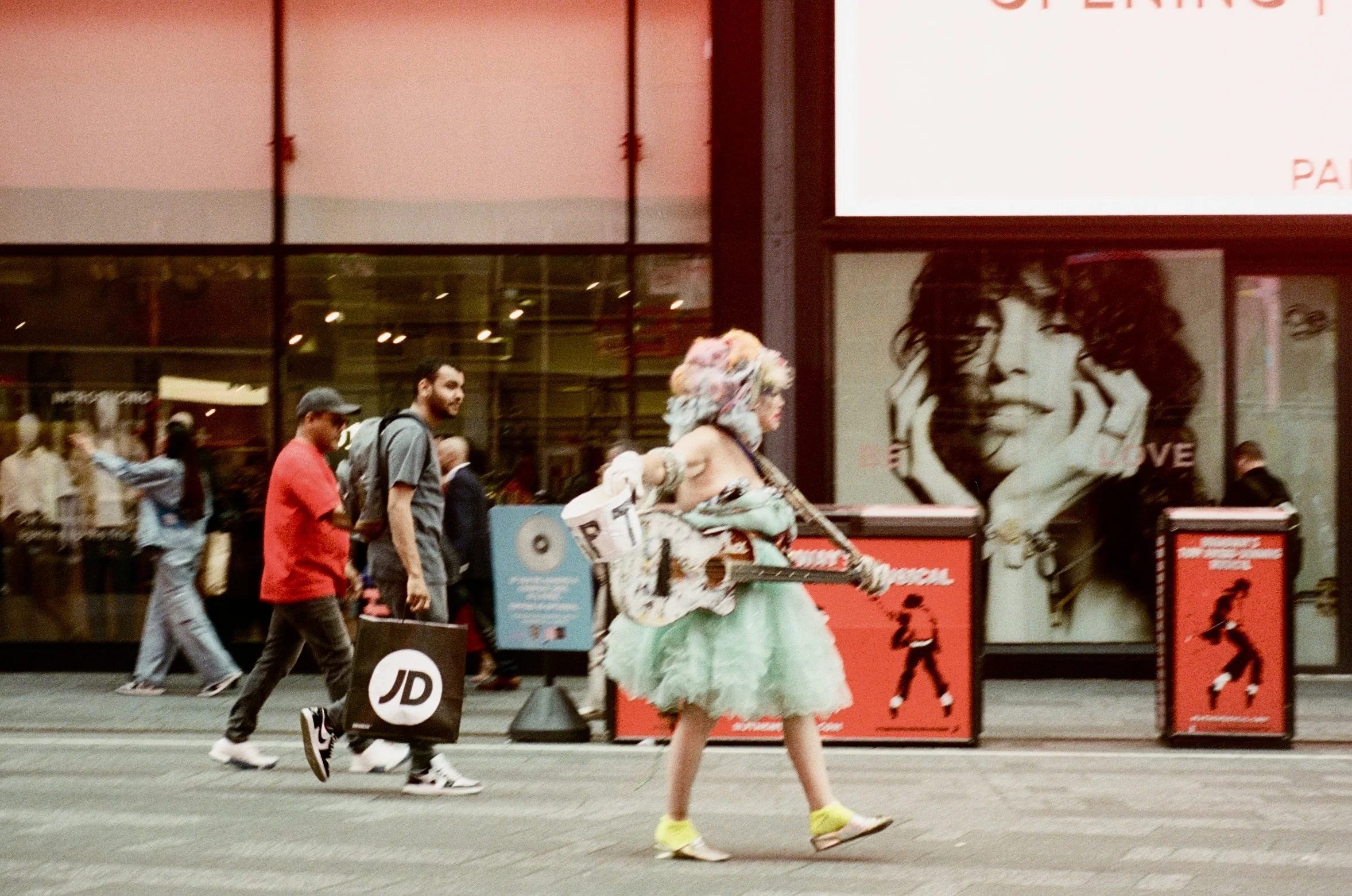 A woman with a colorful, fluffy wig and a green tulle skirt is walking on a city street while holding a guitar and a newspaper. Several pedestrians are walking by in the background, and there are large advertisements on the building behind her.
