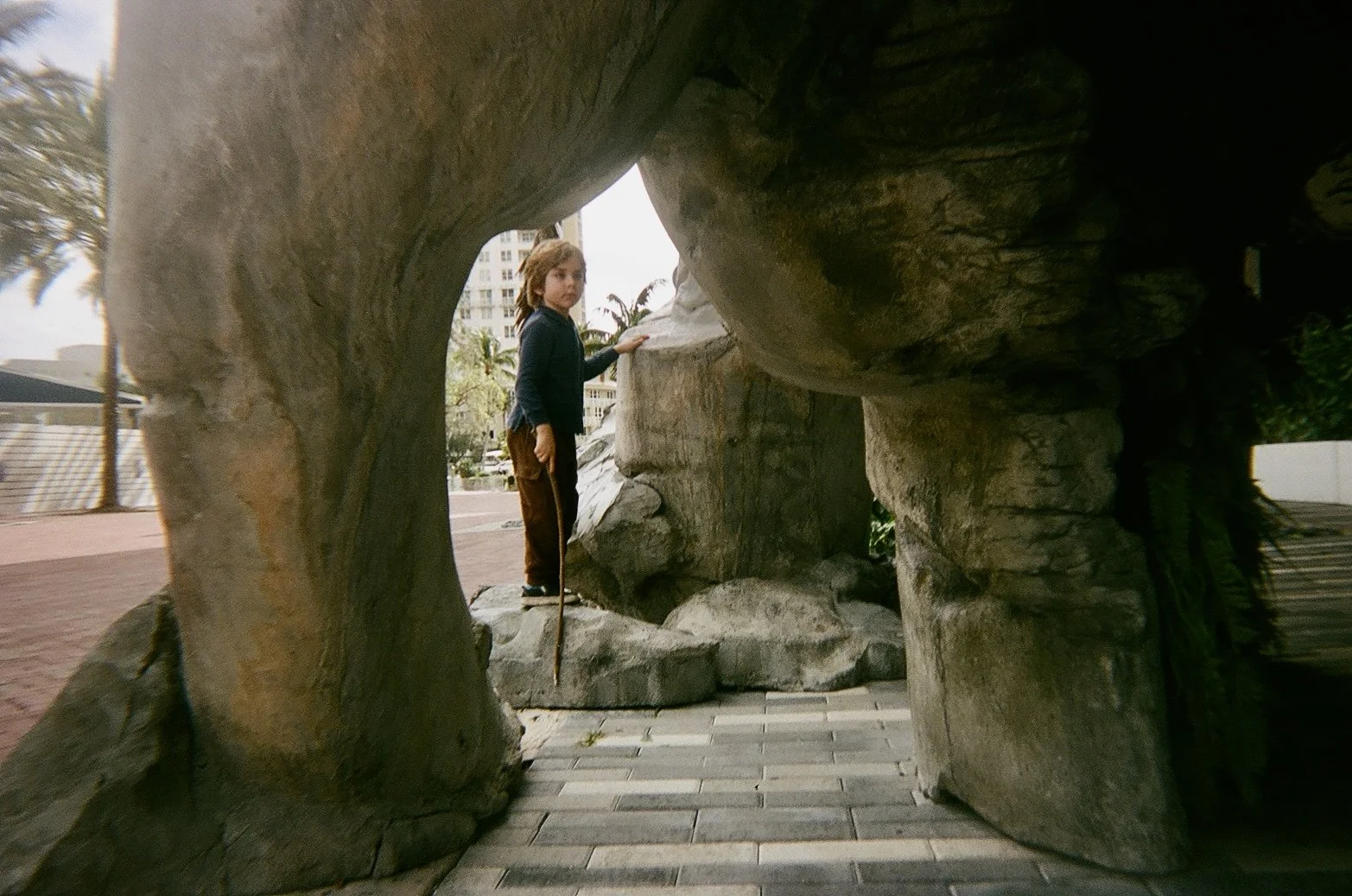 A young boy with long brown hair, wearing a black shirt and brown pants, standing on rocks inside a large rock formation, looking at the camera.
