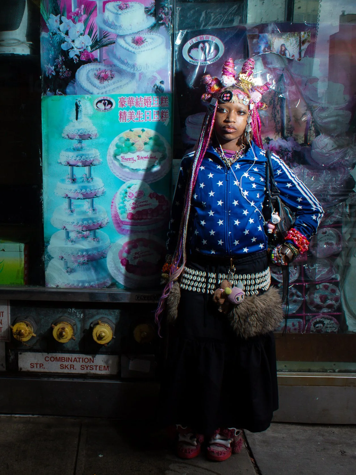 A young woman with elaborate colorful hair ornaments, wearing a star-print blue jacket, standing in front of a cake shop window.