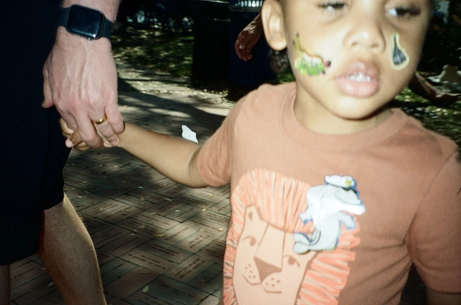 A young child with face paint of a dinosaur and a teardrop sticker on his face, holding hands with an adult, outdoors on a brick pathway with trees in the background.