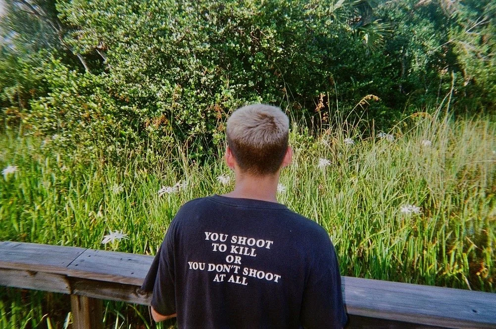 A person with short light hair facing away, wearing a black T-shirt with white text, standing by a wooden railing, overlooking tall grass and bushes in a natural setting.