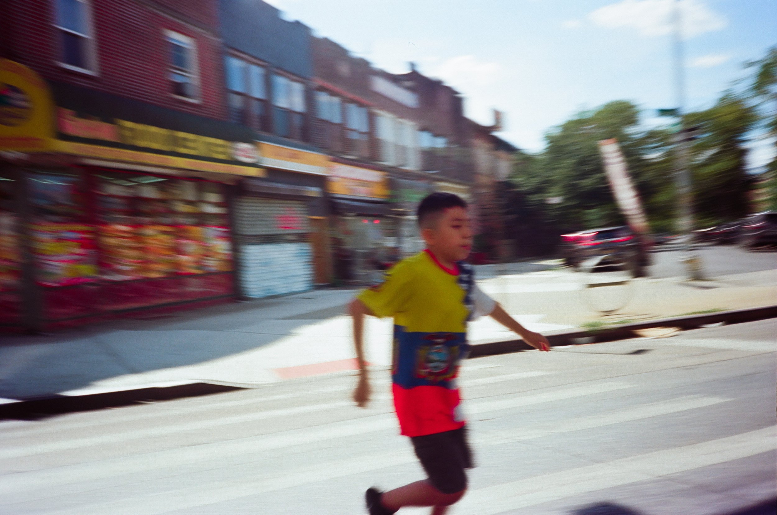 Young boy running across the street in an urban area with storefronts and parked cars in the background.