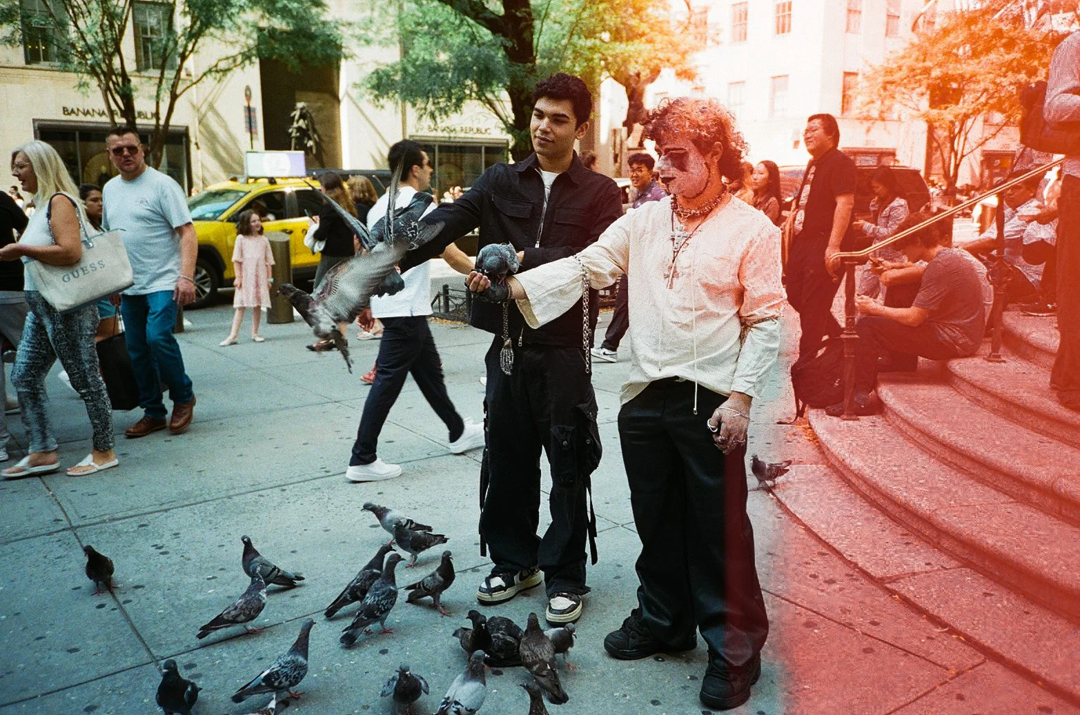 Two men are feeding pigeons on a busy city sidewalk. One man has a skeleton face paint, sunglasses, and gothic style clothing; the other man is young and casually dressed. Several pigeons are on the ground and flying around them. Crowd of pedestrians