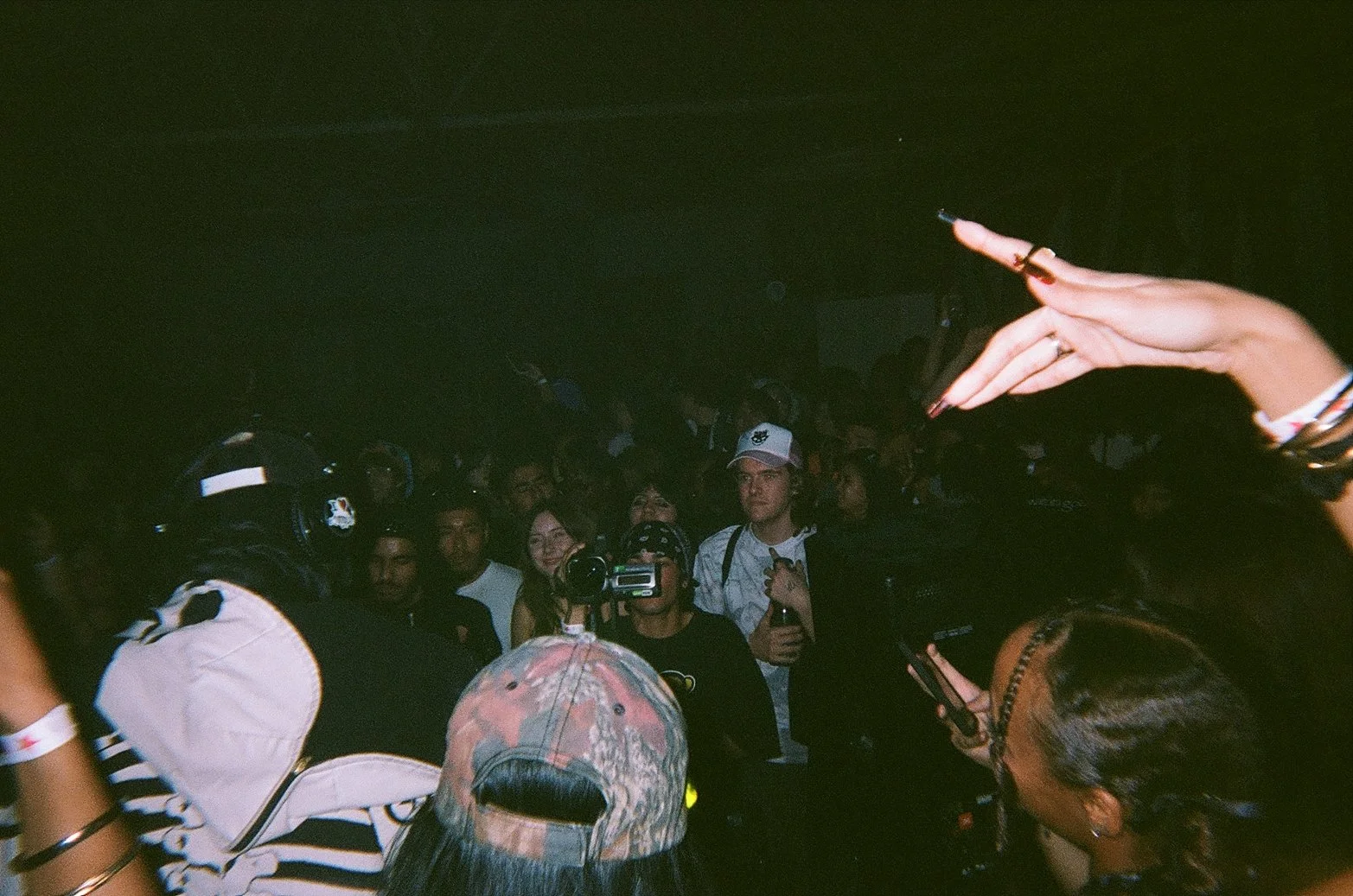 Crowded indoor party with people taking photos and videos, some wearing hats and accessories, dim lighting