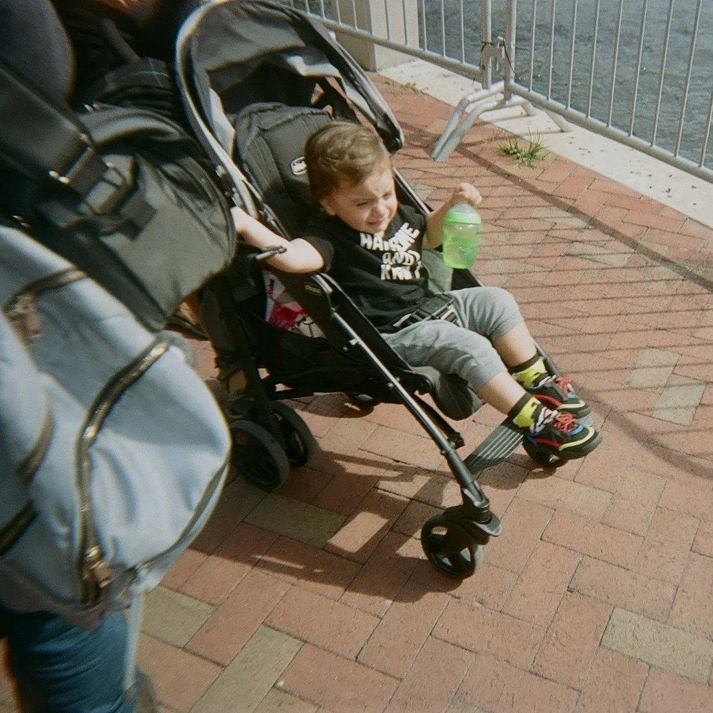 A young boy sitting in a stroller on a brick sidewalk, holding a green bottle, with a metal railing and body of water in the background.