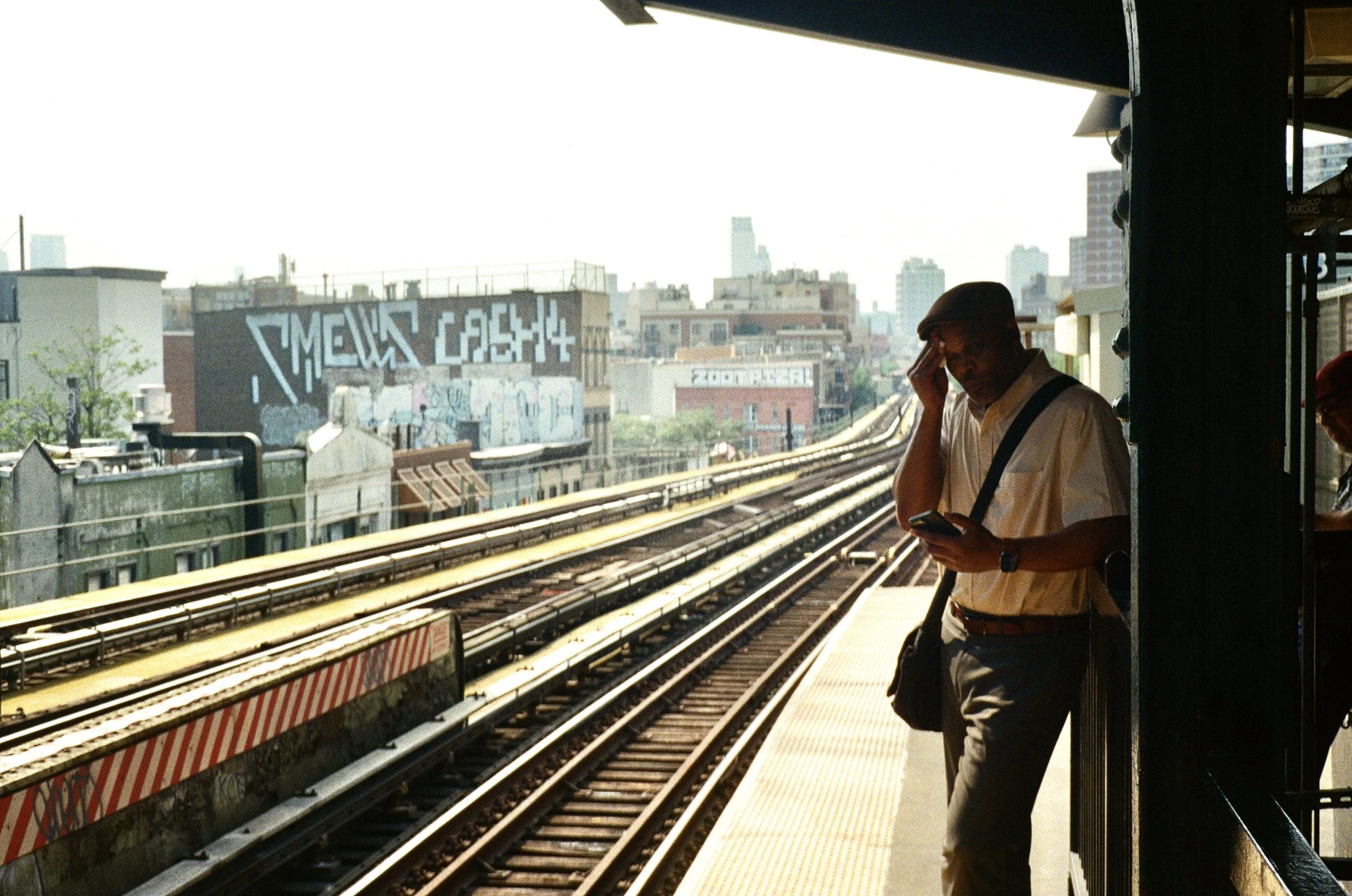 A man in a white shirt and cap standing on a subway platform next to the tracks, looking at his phone with a worried expression, and holding his forehead.