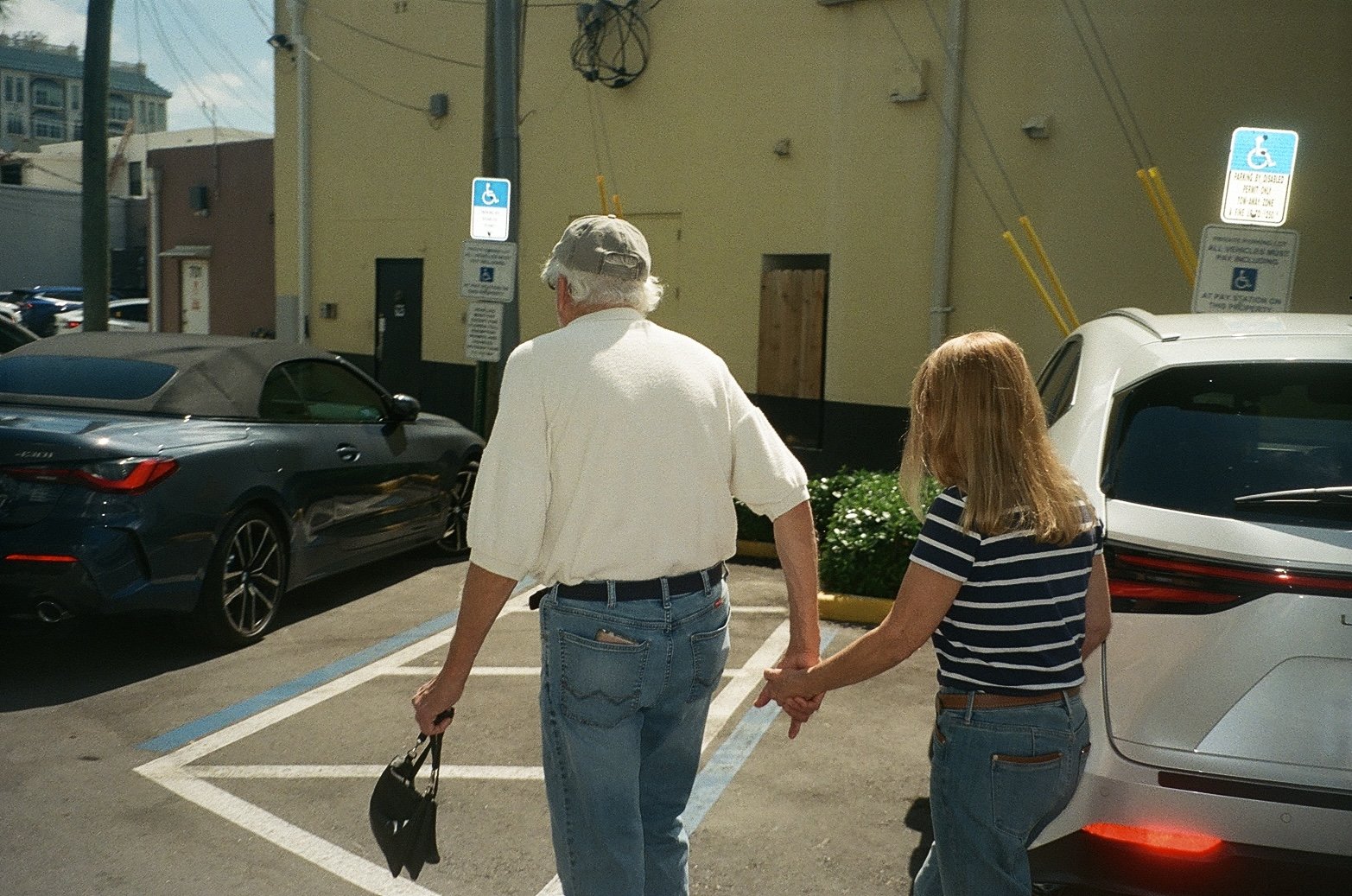 An elderly man and a woman walking hand in hand through a parking lot on a sunny day. The man is wearing a light-colored hat, cream shirt, and jeans, and is holding a black bag. The woman has shoulder-length blonde hair, is wearing a navy and white s