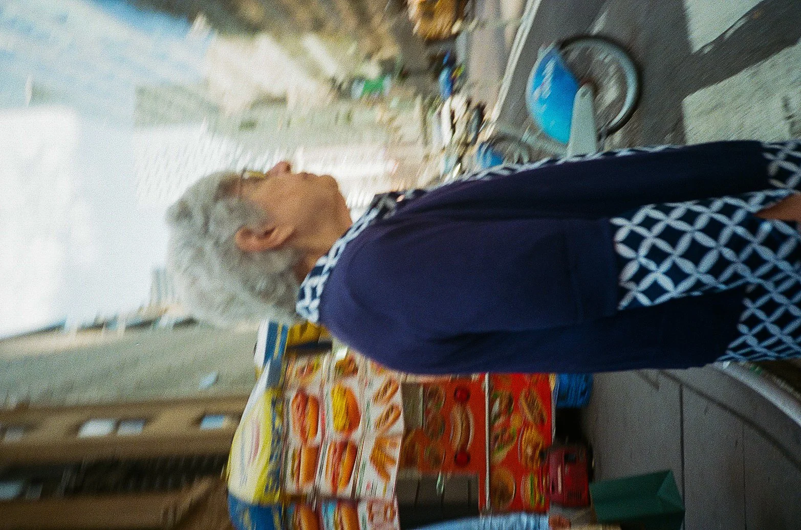Side view of an elderly man with white hair, wearing glasses, a dark jacket with a patterned collar, standing inside a small grocery store with various packaged food items on shelves.