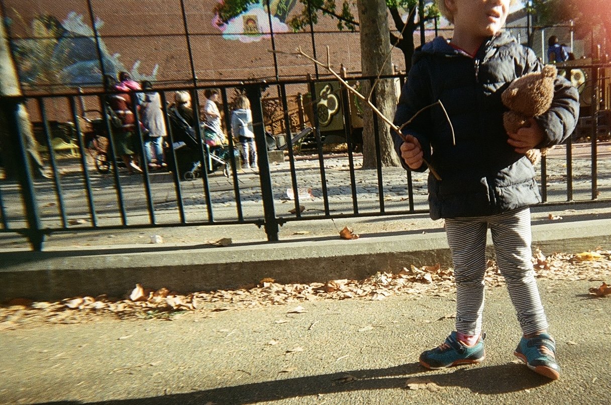 A young child standing on a sidewalk in front of a black metal fence, holding a small stick in one hand and a teddy bear in the other, with playground equipment and people in the background.