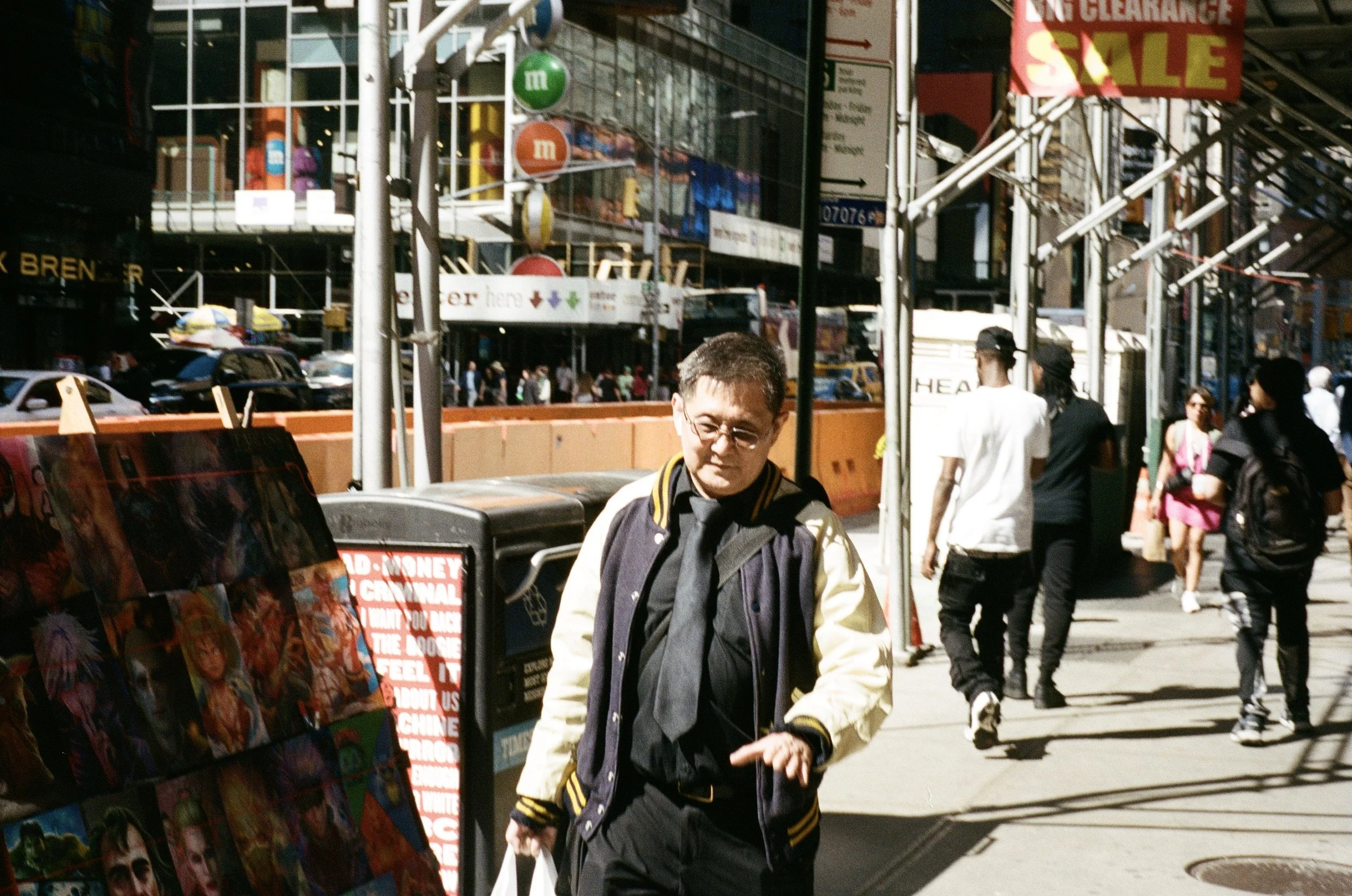 A man in a varsity jacket walking on a busy city sidewalk with shops and people in the background.