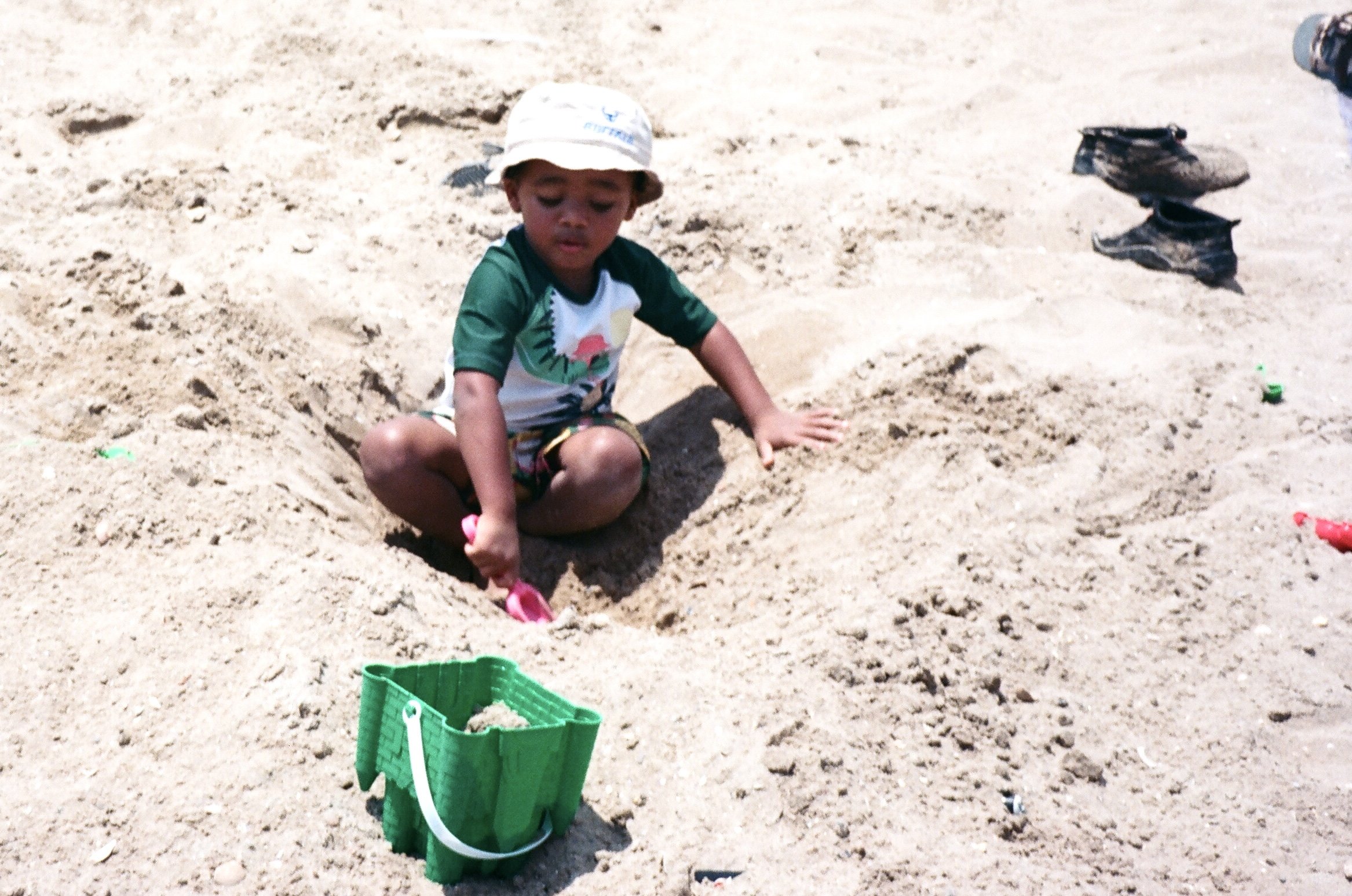 Young boy wearing a hat, green and white shirt, and shorts, playing in the sand at the beach, surrounded by toys and shoes.