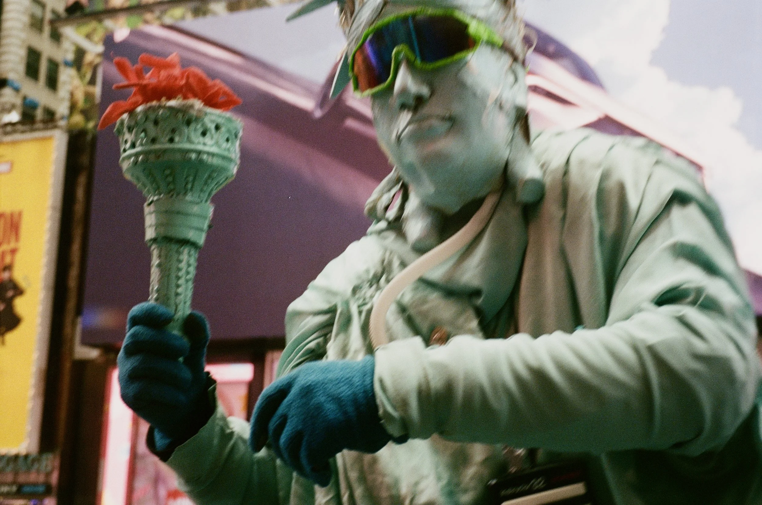 A street performer dressed as a statue, with a metallic body suit and face paint, wearing reflective sunglasses, holding a large decorative torch with red artificial flames, in front of a colorful urban backdrop.