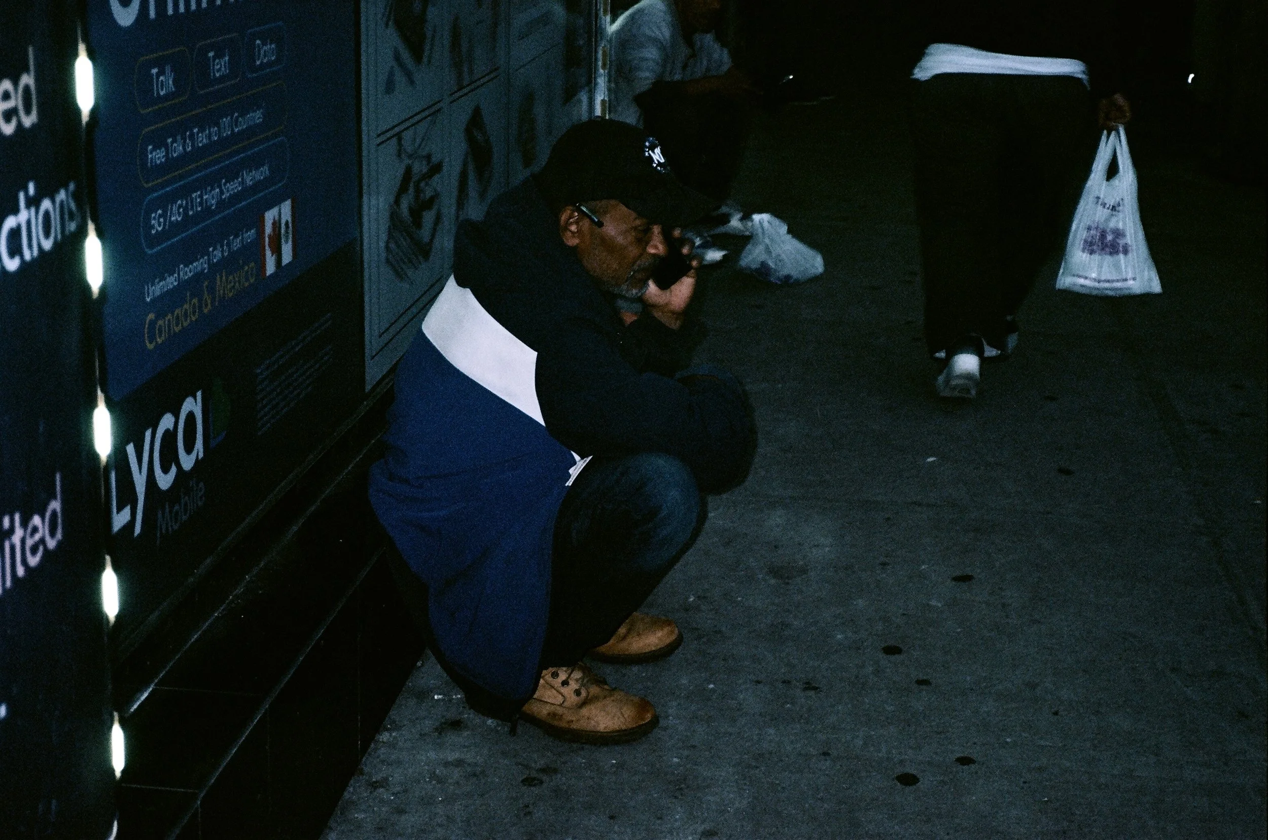 A man crouching on the sidewalk, talking on his phone, wearing a black cap, black jacket with white and blue sections, blue jeans, and brown boots, next to a blue advertisement board.