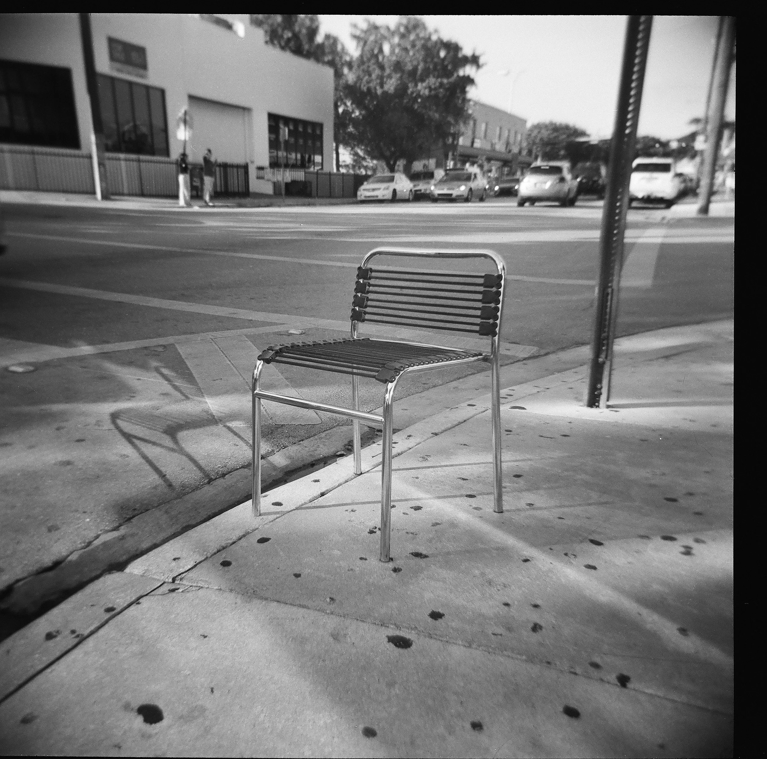 Empty metal bench on sidewalk with street and parked cars in the background, black and white photo.