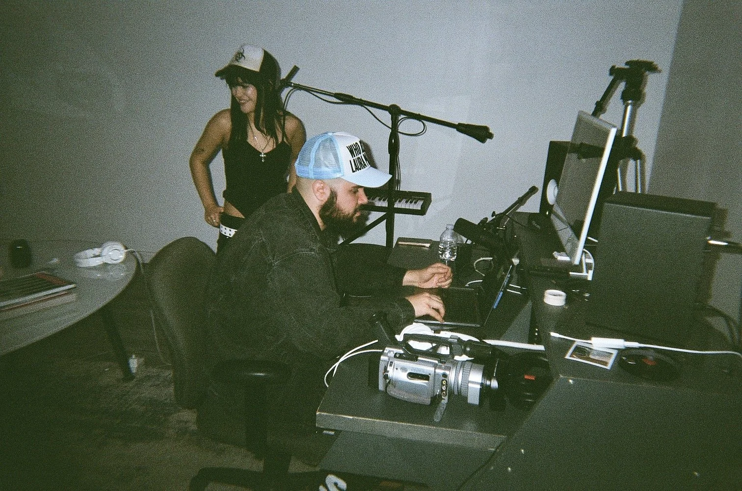 A man wearing a trucker hat with a woman standing behind him in a music studio or editing room with DJ equipment, monitors, and a keyboard.