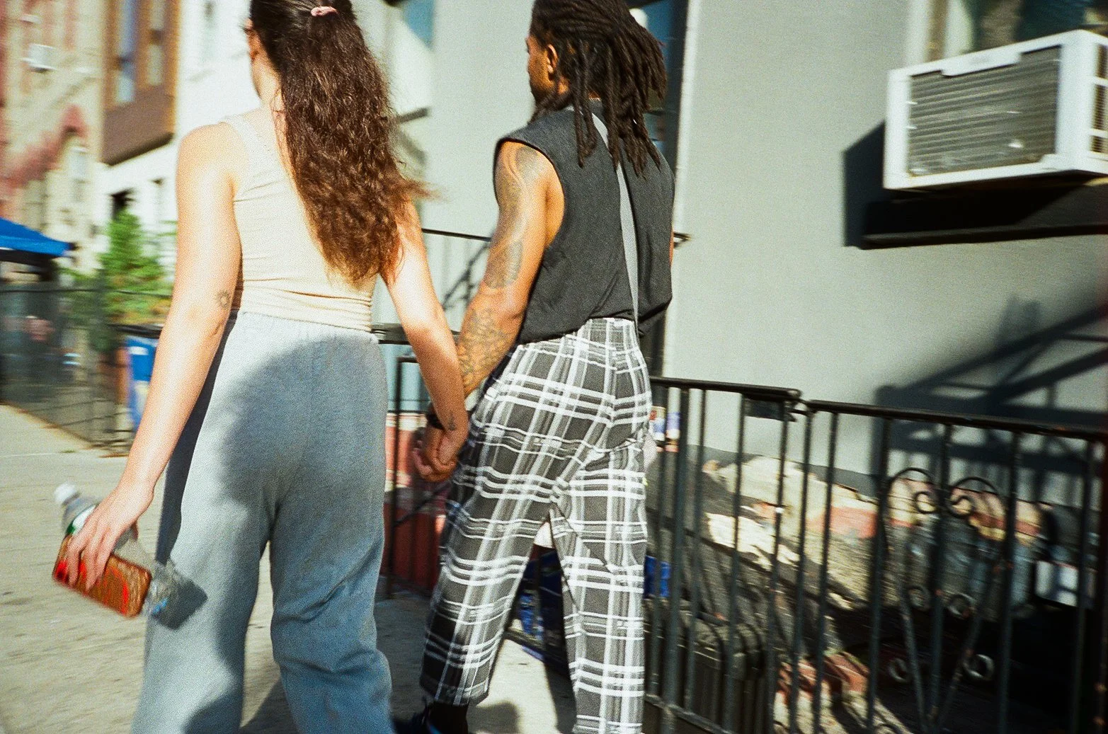 Two people holding hands walking on a city sidewalk. One person has curly hair and is wearing a beige tank top and gray sweatpants, holding a water bottle and a cellphone. The other person has dreadlocks, wearing a black sleeveless shirt and plaid pa