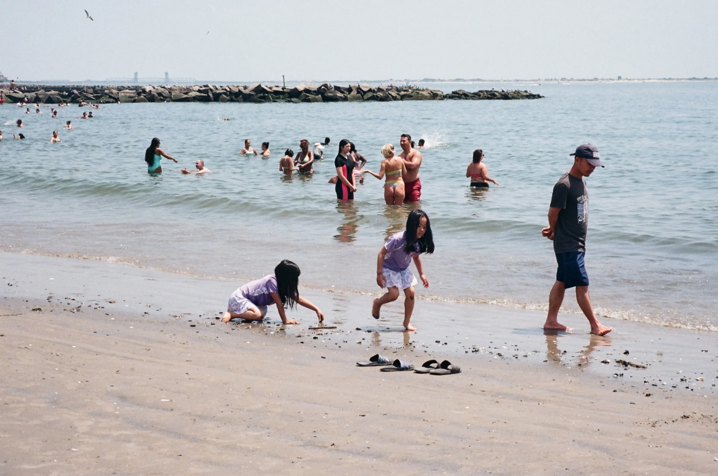 Children playing on the sandy beach near the water, with many people swimming and socializing in the ocean, a rocky breakwater in the background, and seagulls flying overhead on a sunny day.