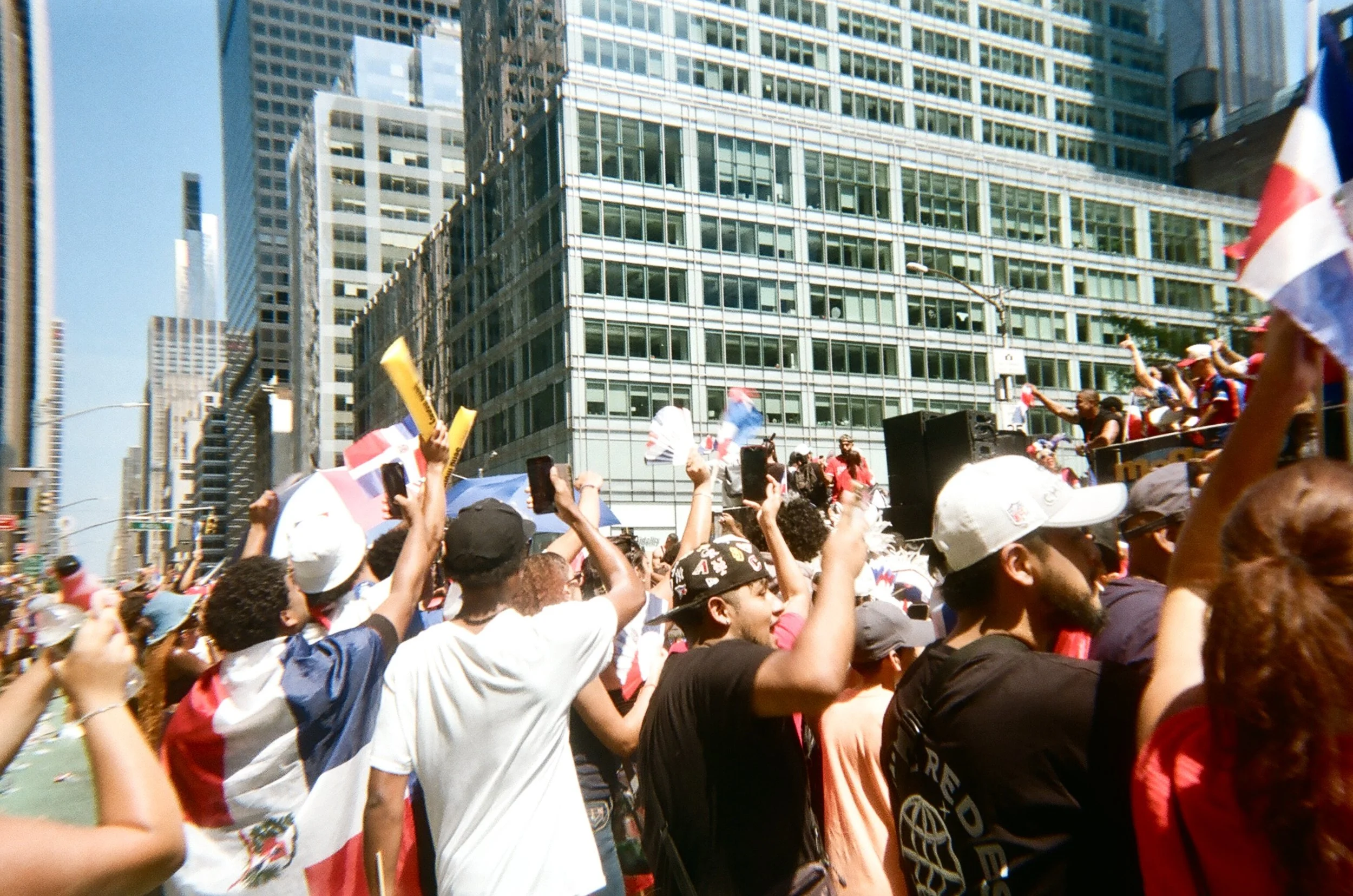 Crowd of people celebrating a parade or protest in an urban area, waving flags and raising their hands, with tall glass buildings in the background on a sunny day.