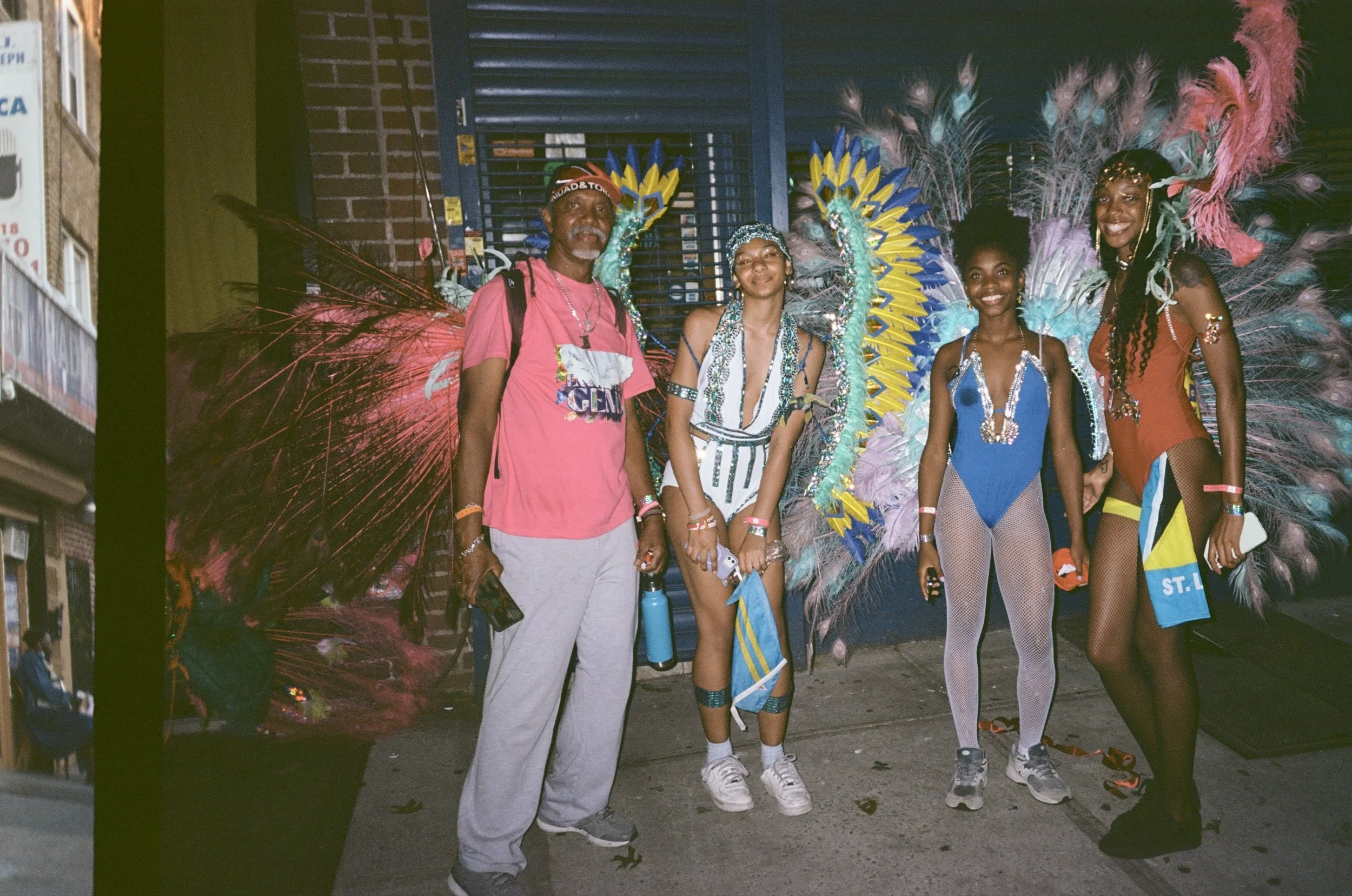 Four people standing in front of a colorful carnival costume with large feathers, smiling at the camera, during a nighttime street event.