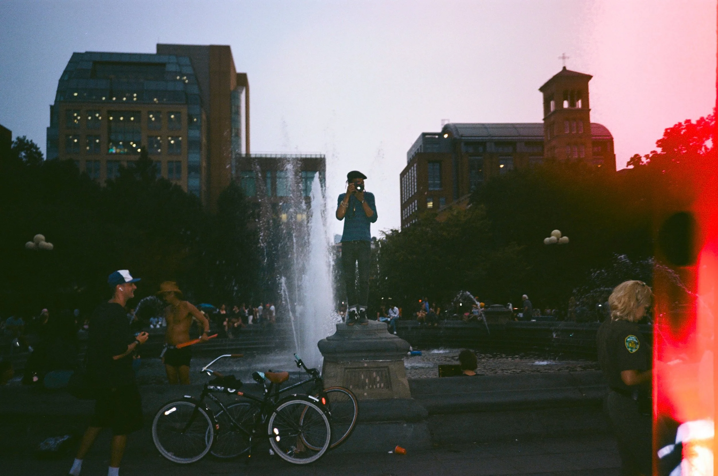 People gathered around a fountain in a city park at dusk, with a person taking a photo on a fountain pedestal and others walking or biking nearby, city buildings in the background.