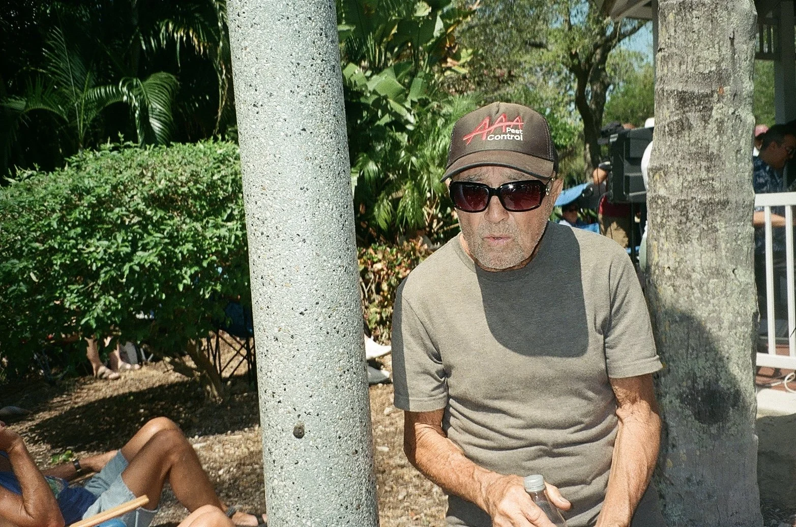 An elderly man wearing sunglasses, a brown baseball cap, and a gray t-shirt, standing outdoors between two palm trees with lush green foliage in the background.