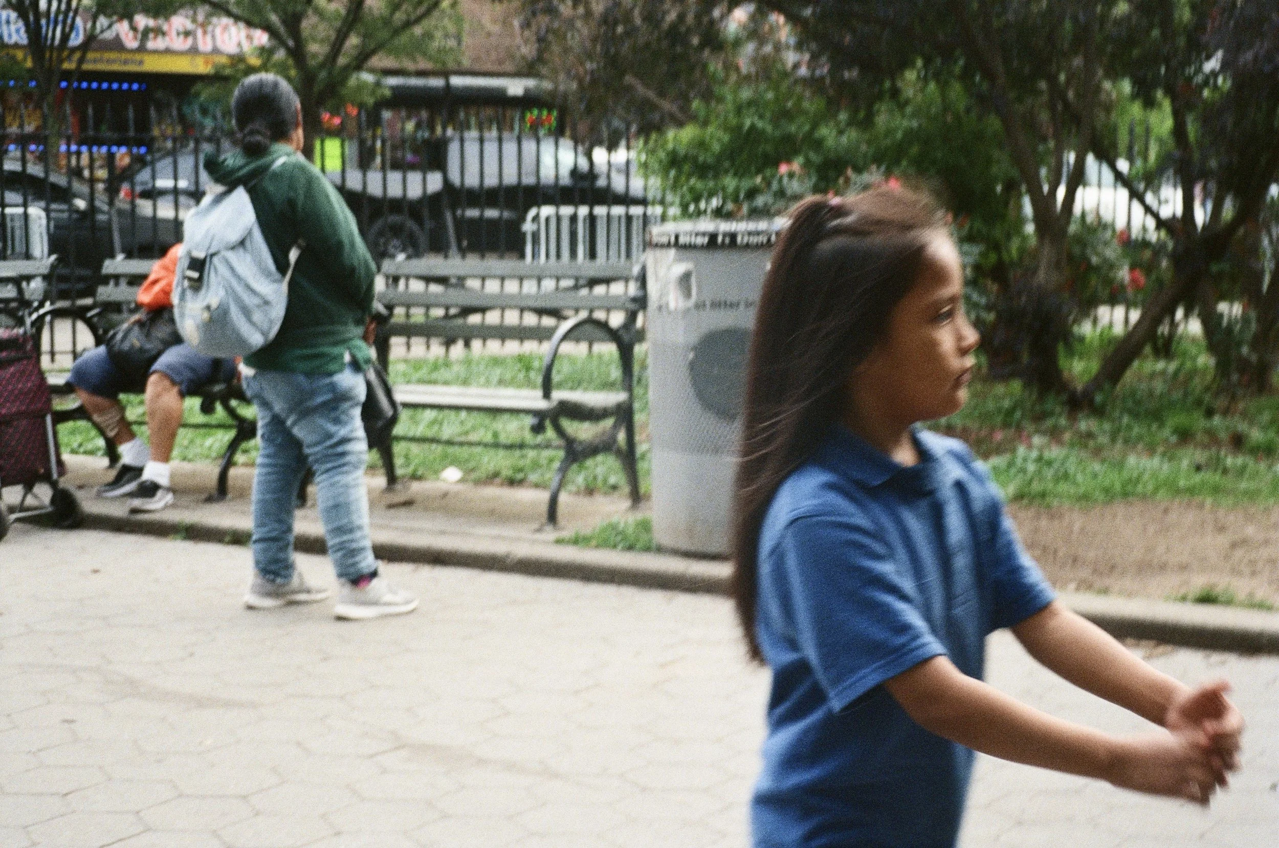 A young girl in a blue shirt is running or playing outside in a park with trees and benches. In the background, a woman with a backpack stands near a bench, and a person is seated with legs stretched out, possibly resting.