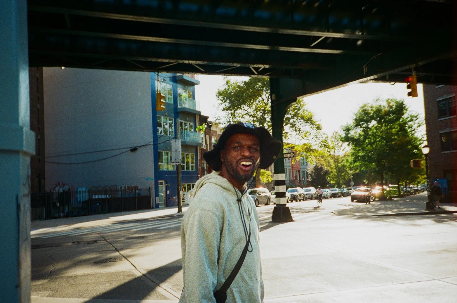 A man smiling and looking at the camera under an overpass on a city street, with trees and parked cars in the background during daylight.