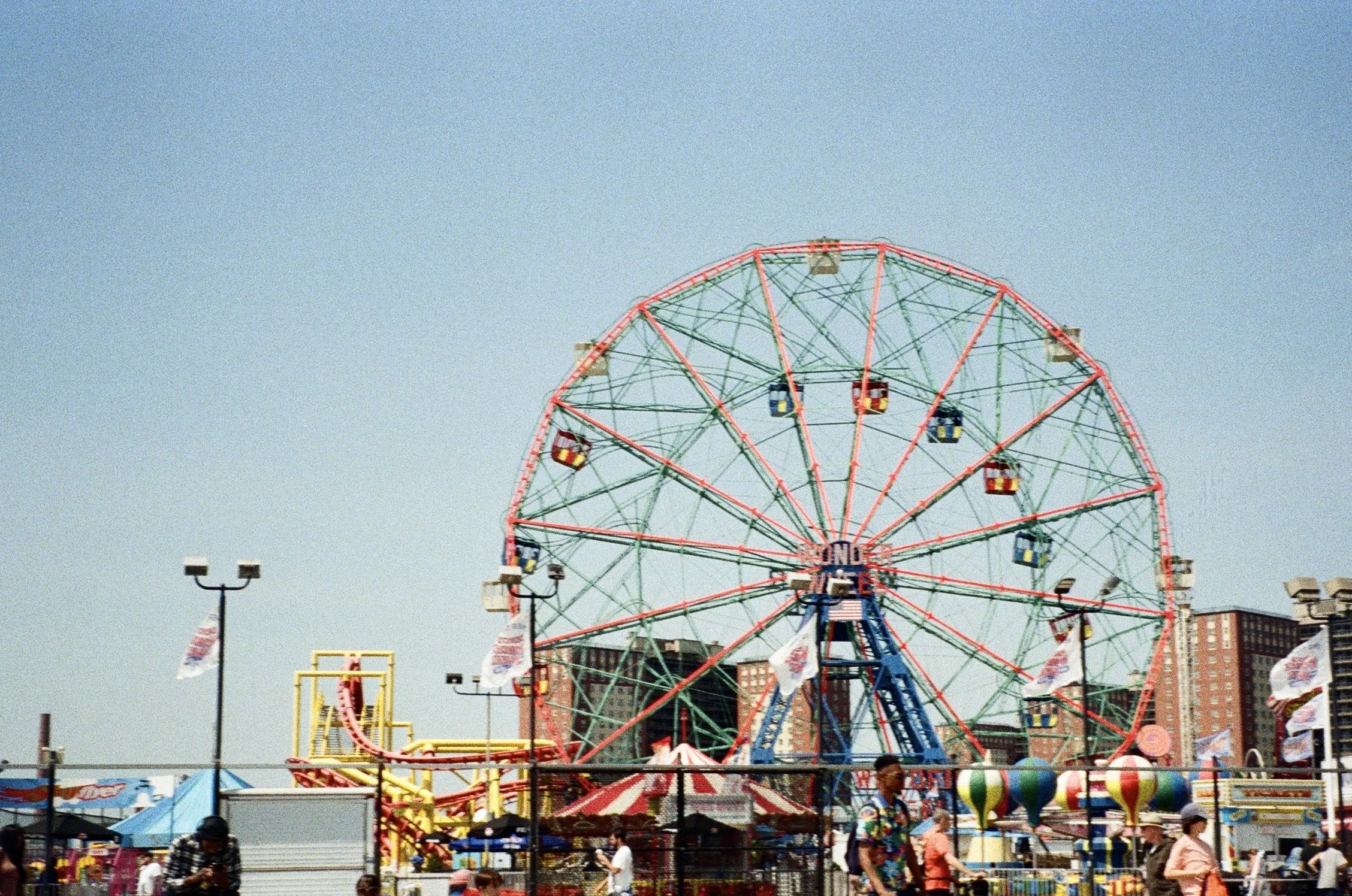 A large amusement park Ferris wheel with colorful passenger cabins, set against a clear blue sky, with several people walking around and various carnival rides and stalls visible in the foreground.