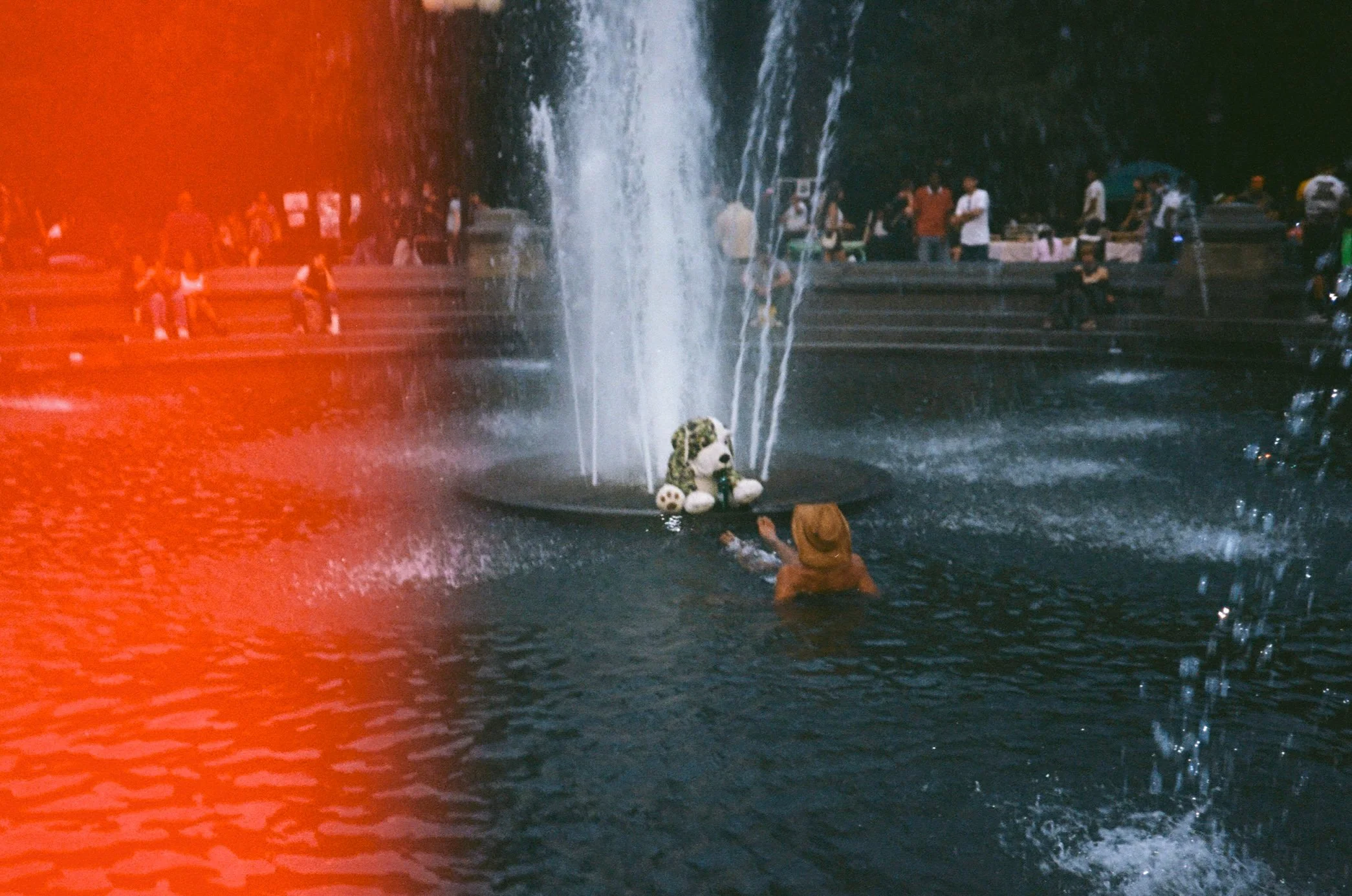 People gathered around a fountain with a panda sculpture and soccer balls, water shooting from the fountain, some watching from the steps, and a person in a hat swimming in the pool.