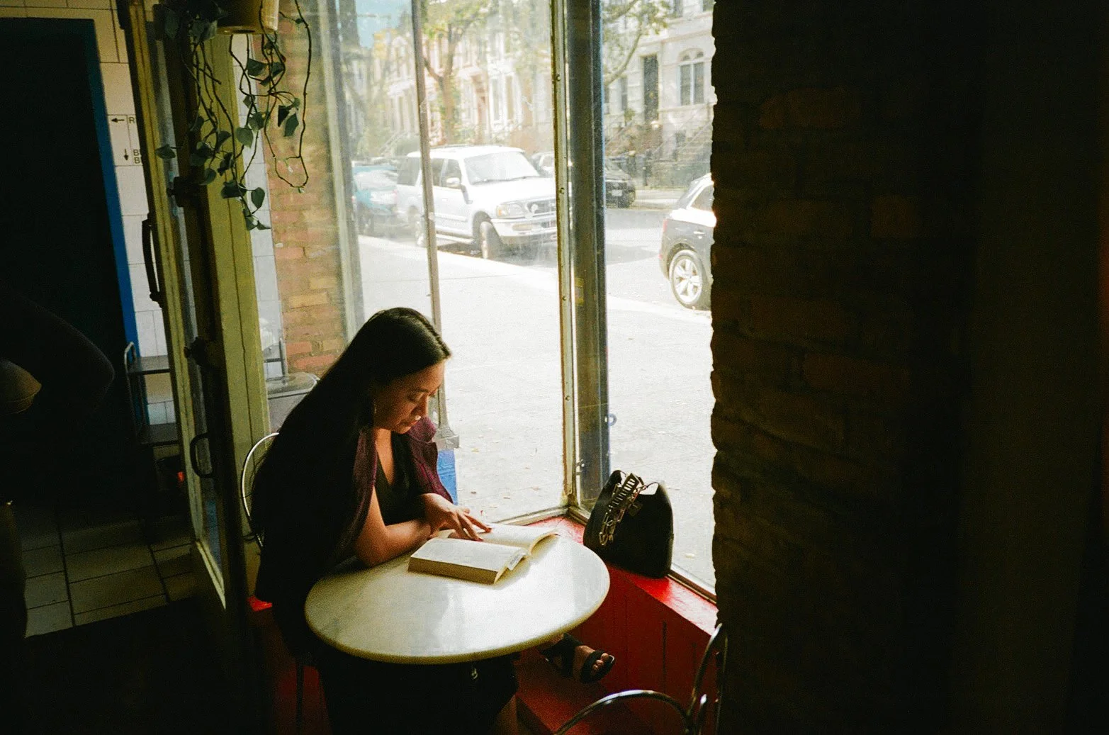 A woman reading a book at a small round table near a large window in a cafe, with cars visible on the street outside.