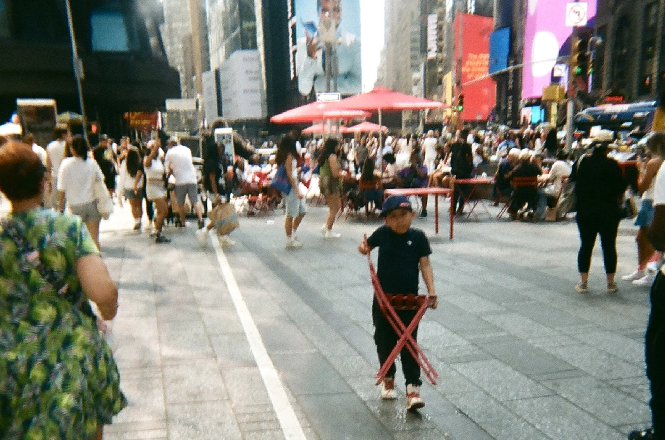 A busy city street with many pedestrians, some sitting at outdoor tables under red umbrellas. A young child in black clothing and a cap is walking across the street carrying a red foldable chair. Tall buildings and electronic billboards are visible i