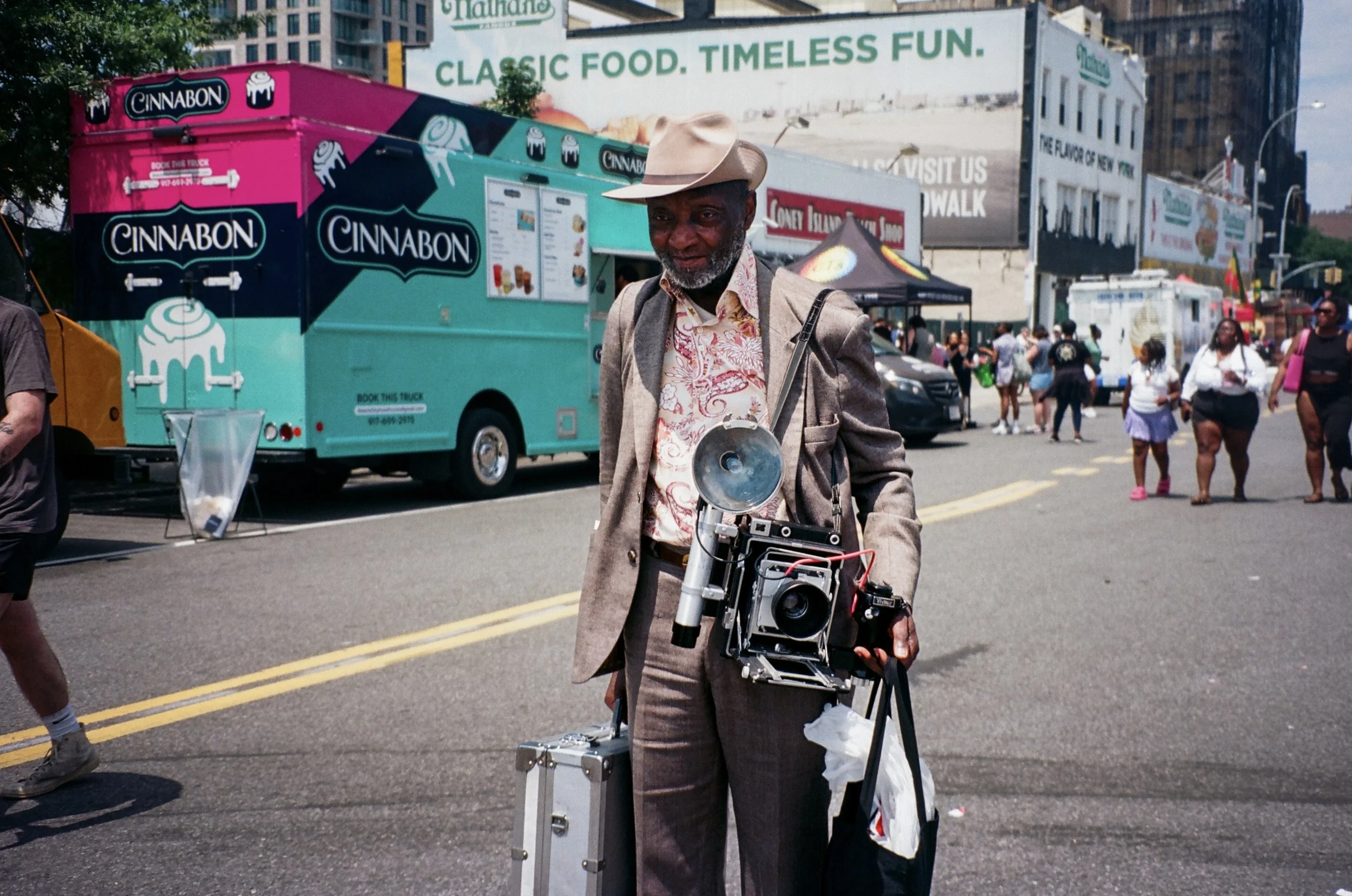 An older man in a light brown suit and hat carrying a vintage camera around his neck, holding a metal case and a black tote bag on a city street with food trucks, tents, and pedestrians in the background.