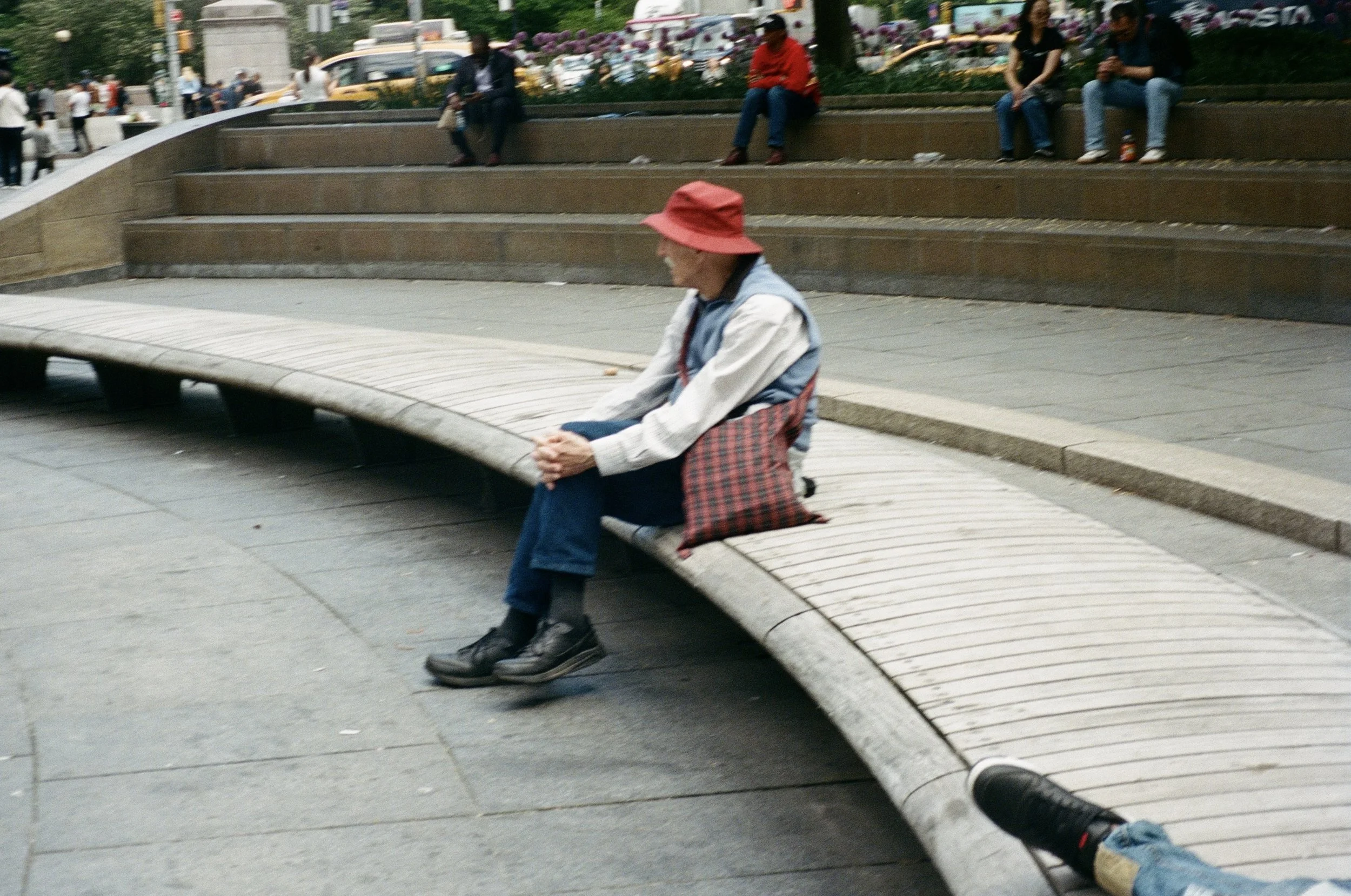 A man sitting alone on a curved wooden bench in an urban park, wearing a red hat, blue vest, and dark shoes, with a plaid bag beside him. In the background, several people are sitting and standing, with cars and trees visible behind them.