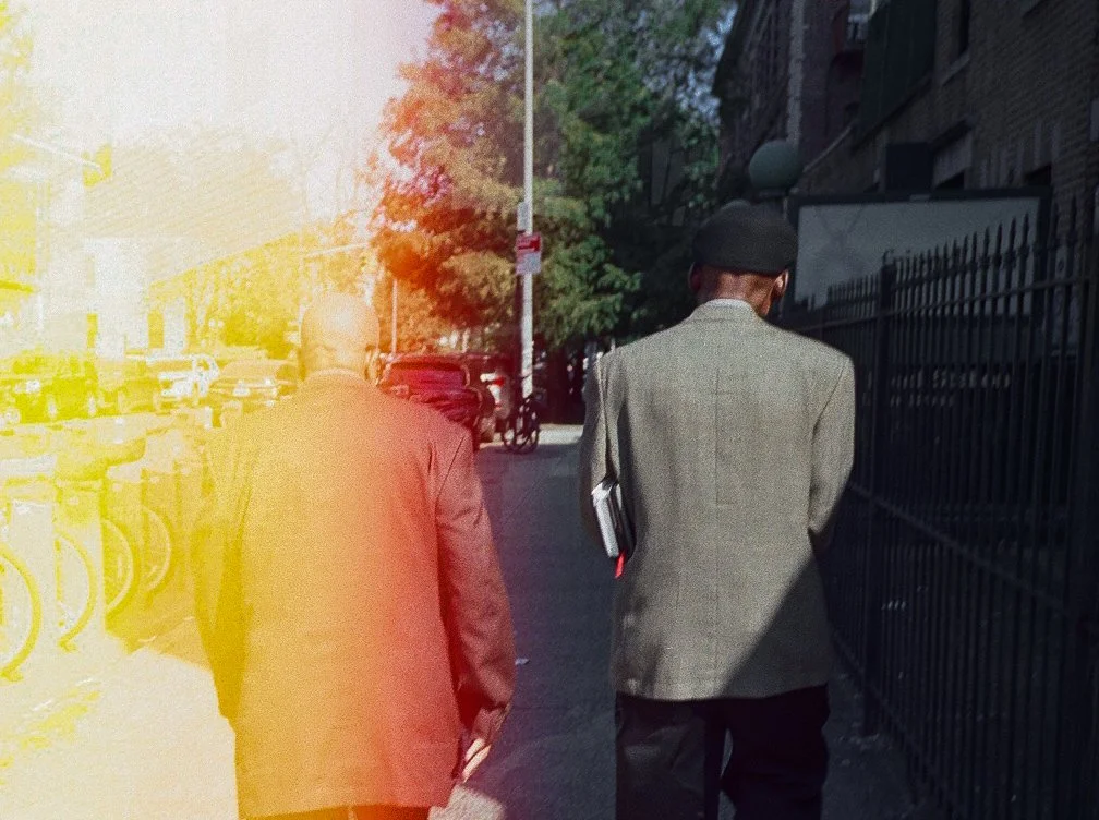 Two men in suits walking down city sidewalk with parked bikes and cars, trees, and buildings in the background.