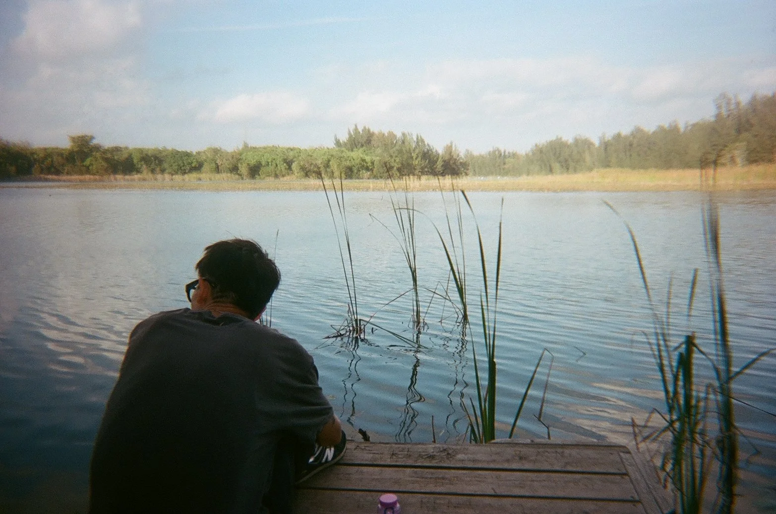 A person with dark hair and glasses sitting on a wooden dock by a calm lake, with tall grass in the water and trees in the distance under a partly cloudy sky.